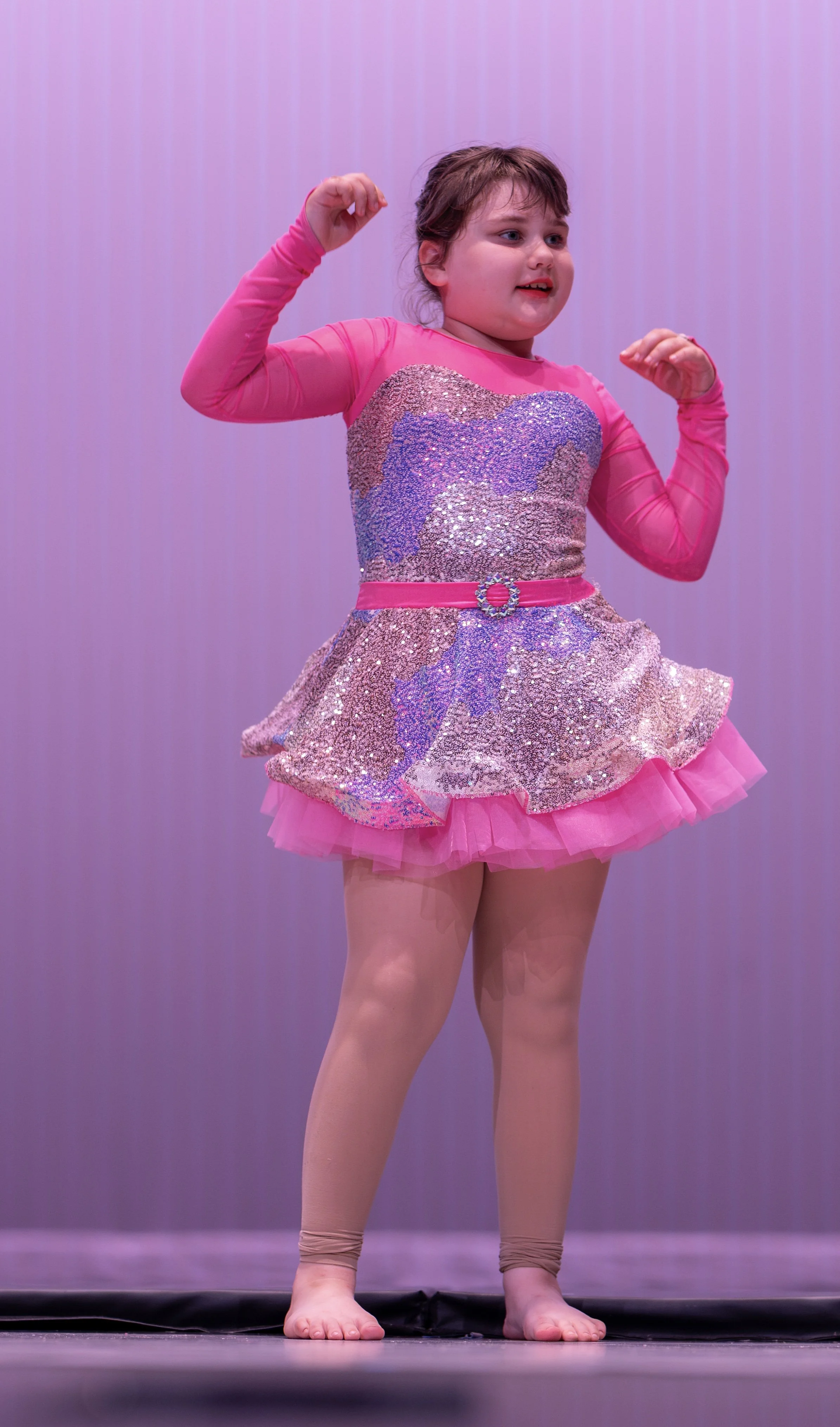 Young girl performing a dance on stage, wearing a sparkly purple and pink tutu with a pink belt, long pink sleeves, tan legs, and pink leggings.