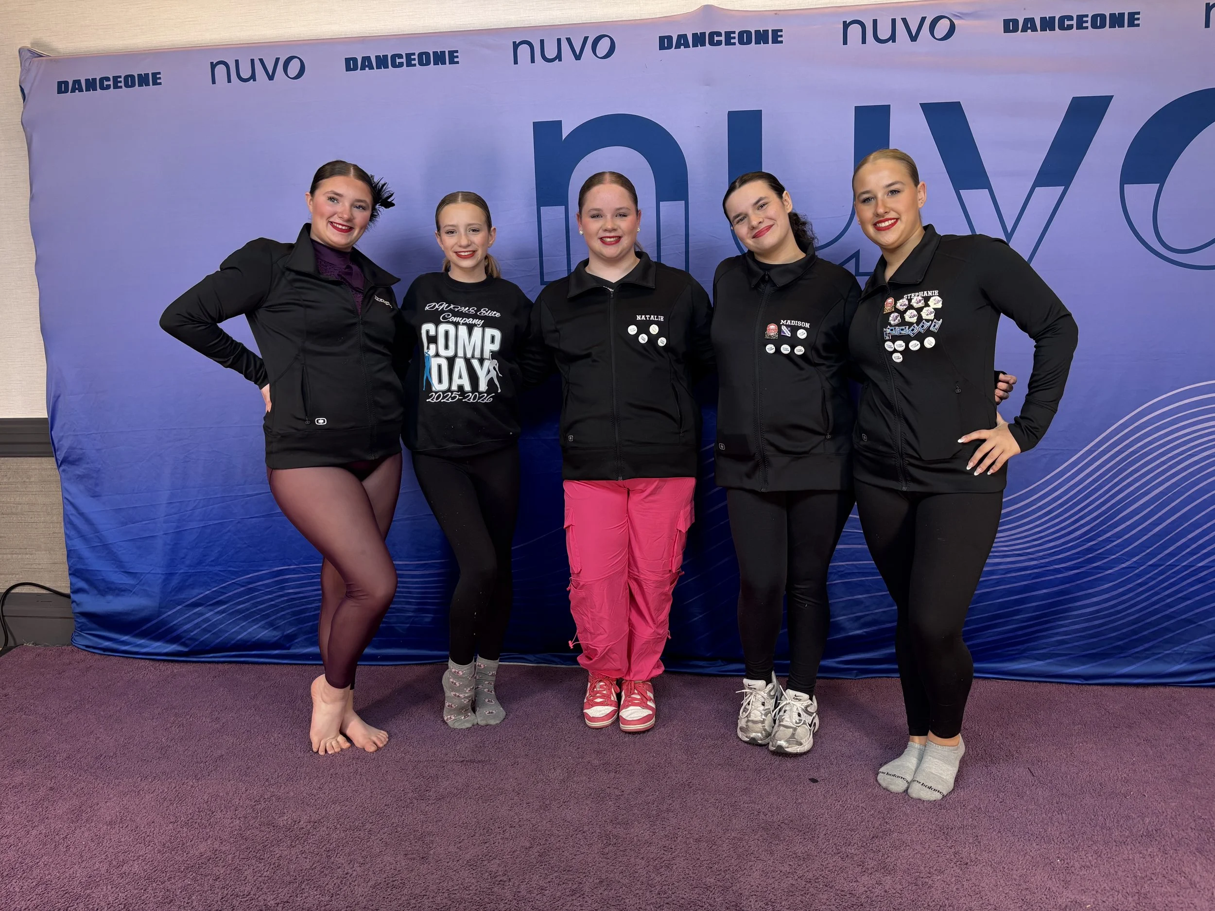 Five young female gymnasts standing side by side in front of a blue and purple backdrop with the words "NUVO" and "DANCEONE" repeated on it. They are wearing black jackets, with some having medals and badges on them, and all are smiling at the camera