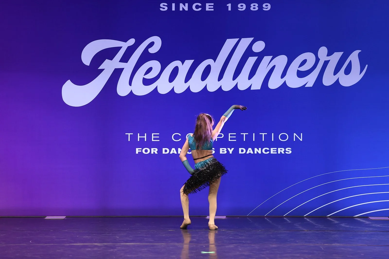 A dancer performing on stage in front of a large purple banner with 'Headliners' and 'Since 1989' written on it, dressed in a blue and black costume, raising her right arm.