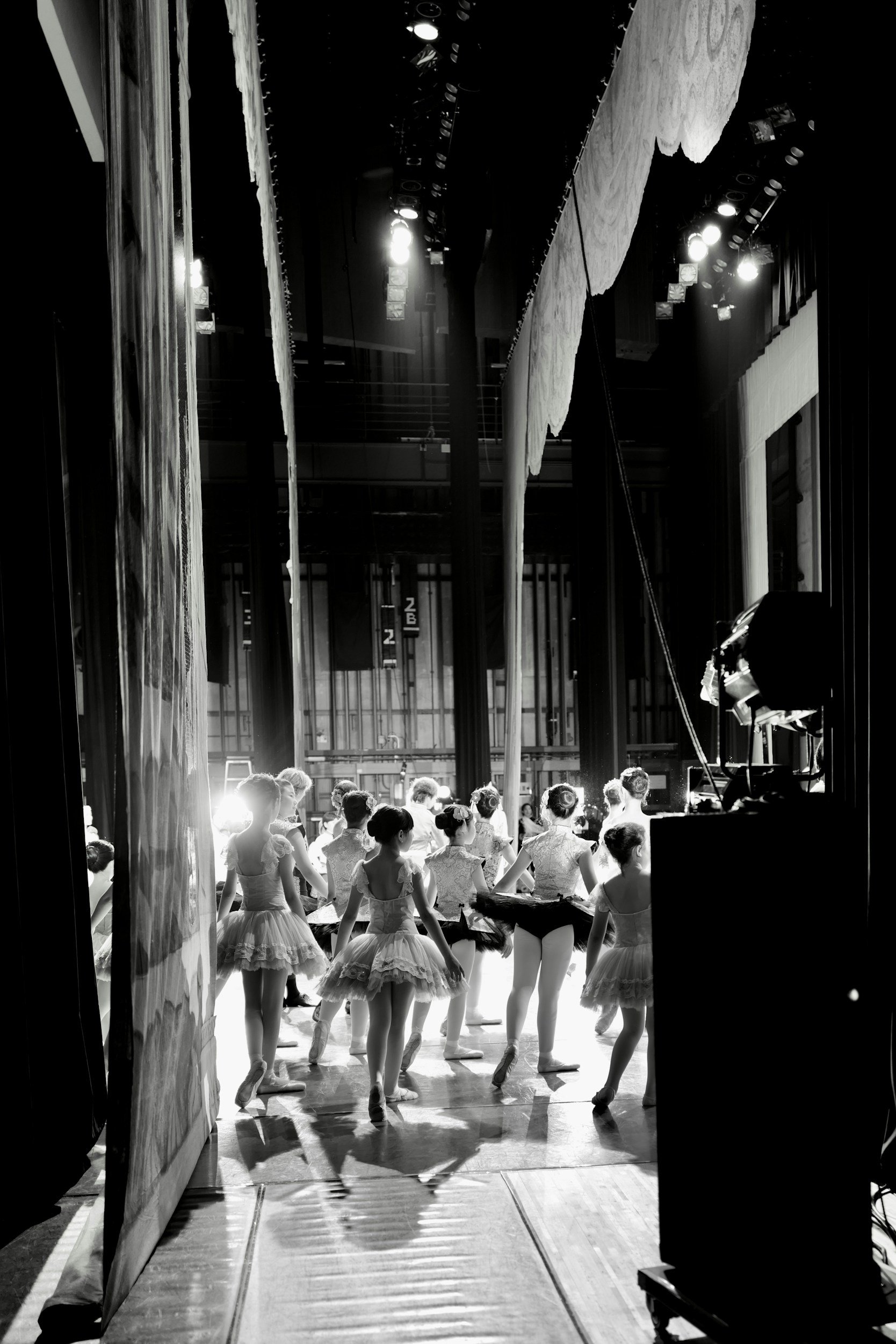 Ballet dancers backstage preparing to perform, seen from behind through a partially open curtain, with stage lighting illuminating them.