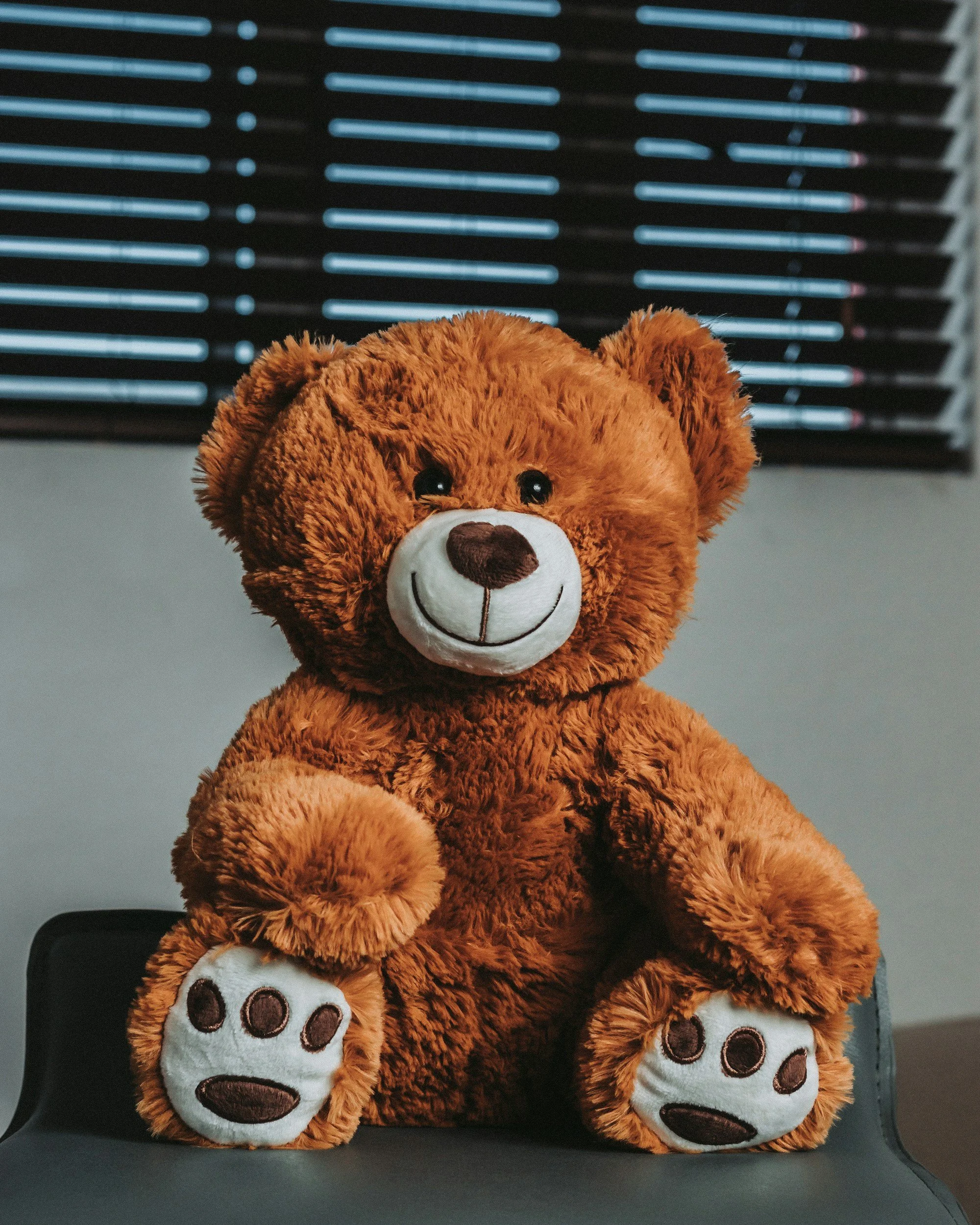 A brown teddy bear with a white snout, smiling face, and paw prints on its paws, sitting on a black surface with a blue and black striped background.