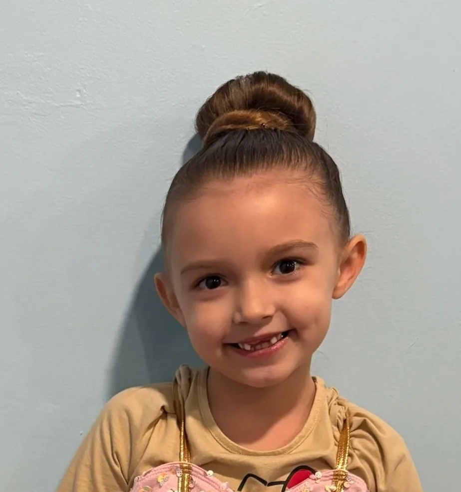 Young girl with a high bun hairstyle, smiling, wearing a beige shirt, holding a pink plush toy.