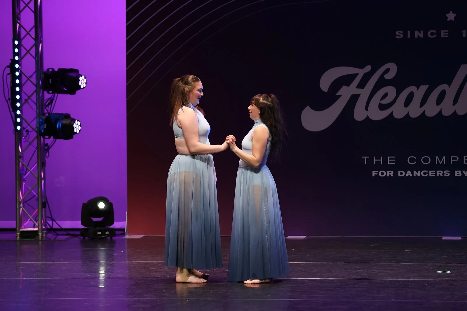 Two female dancers holding hands and looking at each other on stage, dressed in light blue dance costumes with sheer skirts.