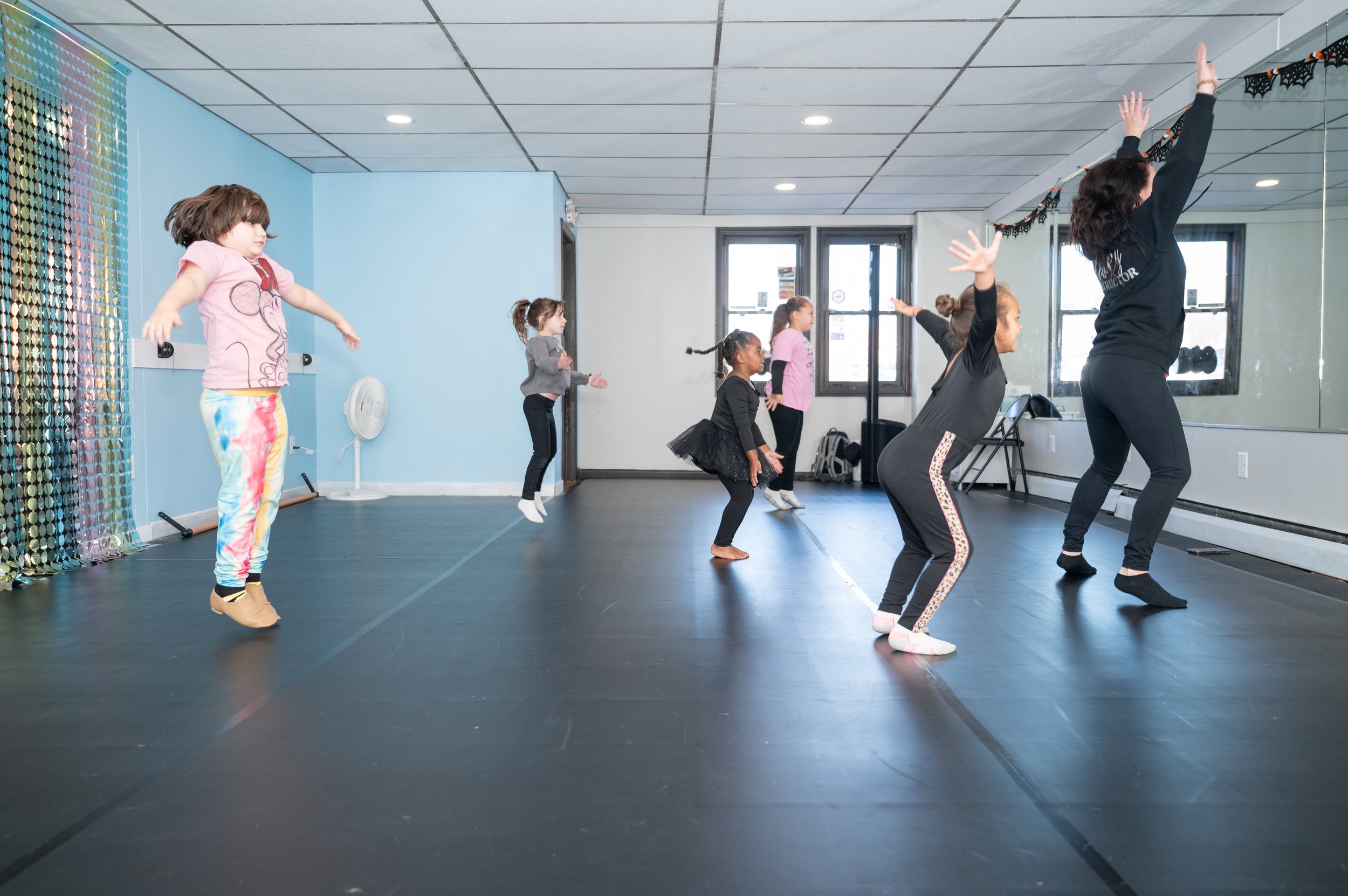 Young girls and an instructor practicing dance moves in a dance studio with black flooring, blue and white walls, and a large mirror. The girls are jumping or stretching, and the instructor is demonstrating a pose with arms raised.