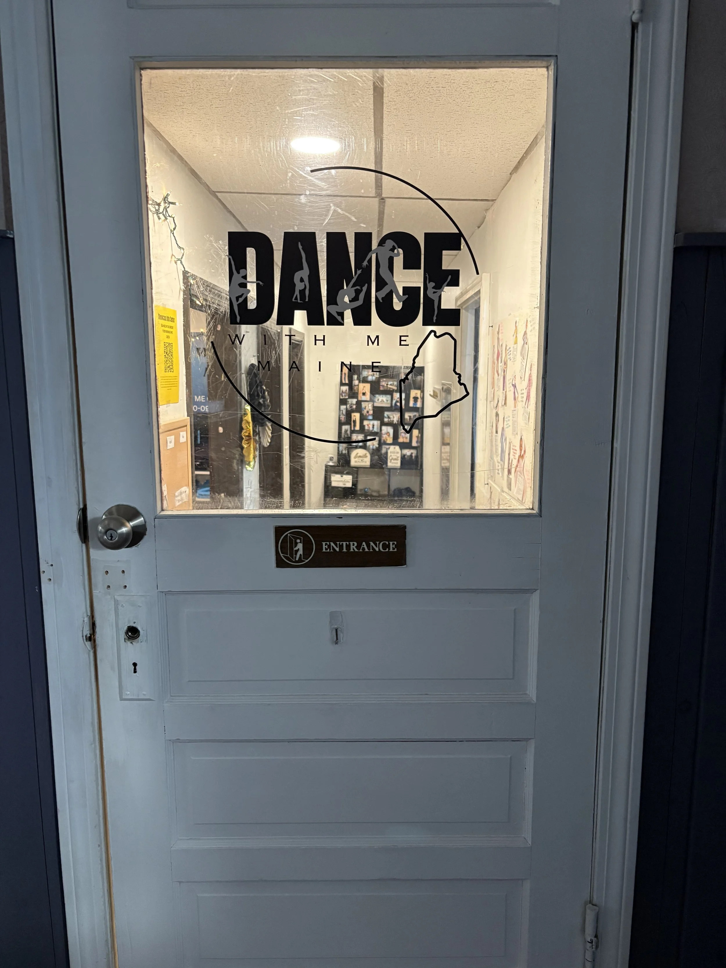 White door with a glass window that has a 'DANCE WITH ME MAINE' sticker featuring silhouettes of people dancing and an outline of Maine on the glass. There is a brown 'ENTRANCE' sign below the window. The door has a silver doorknob and a keyhole.