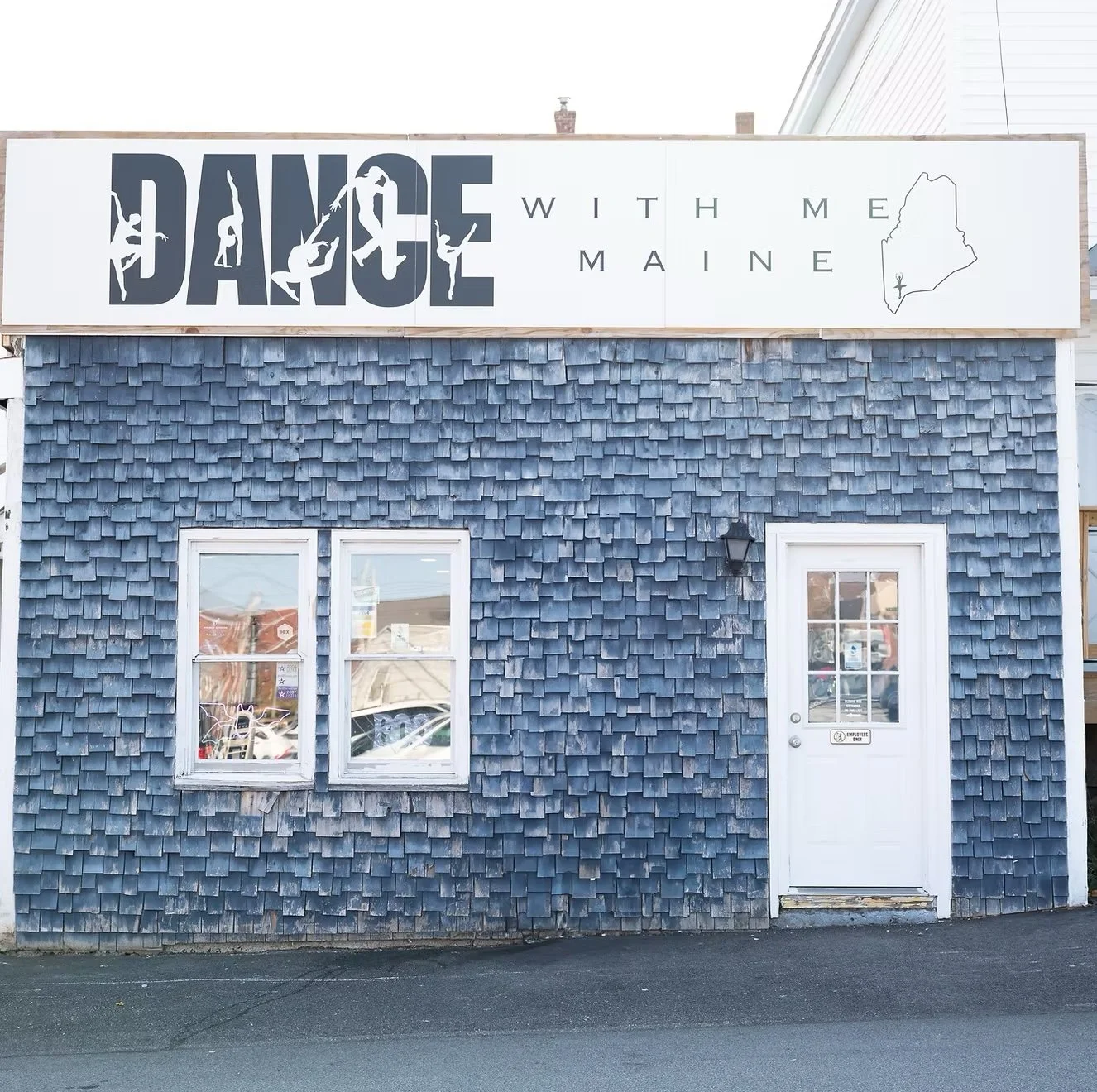 Blue building with shingle siding and a white door, with a sign on top reading 'Dance with Me Maine' and an outline of the state of Maine.