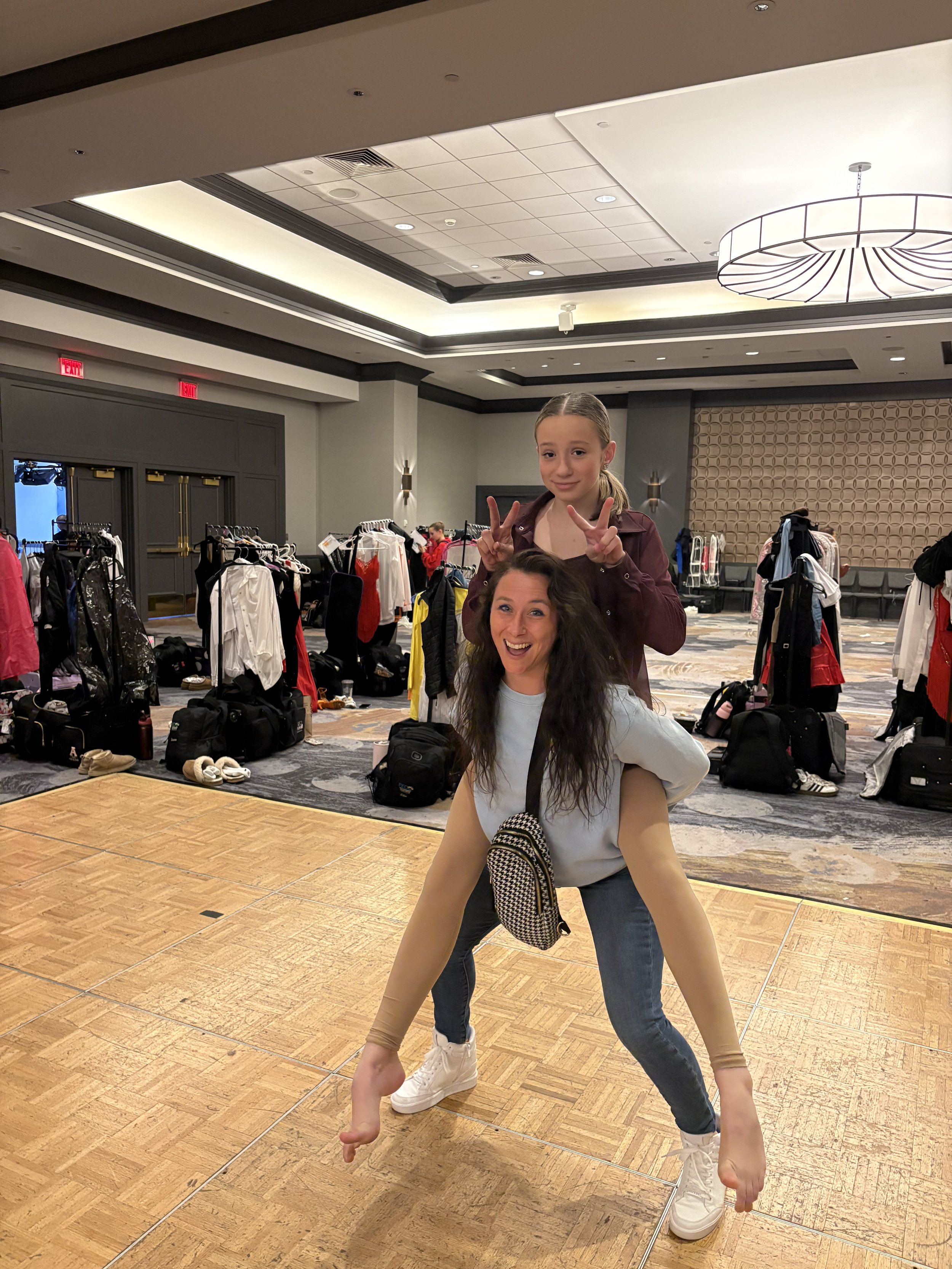 Two women, one older and one younger, posing playfully in an indoor space with clothing racks and bags in the background. The younger woman is sitting on the older woman's shoulders, and both are making peace signs and smiling.