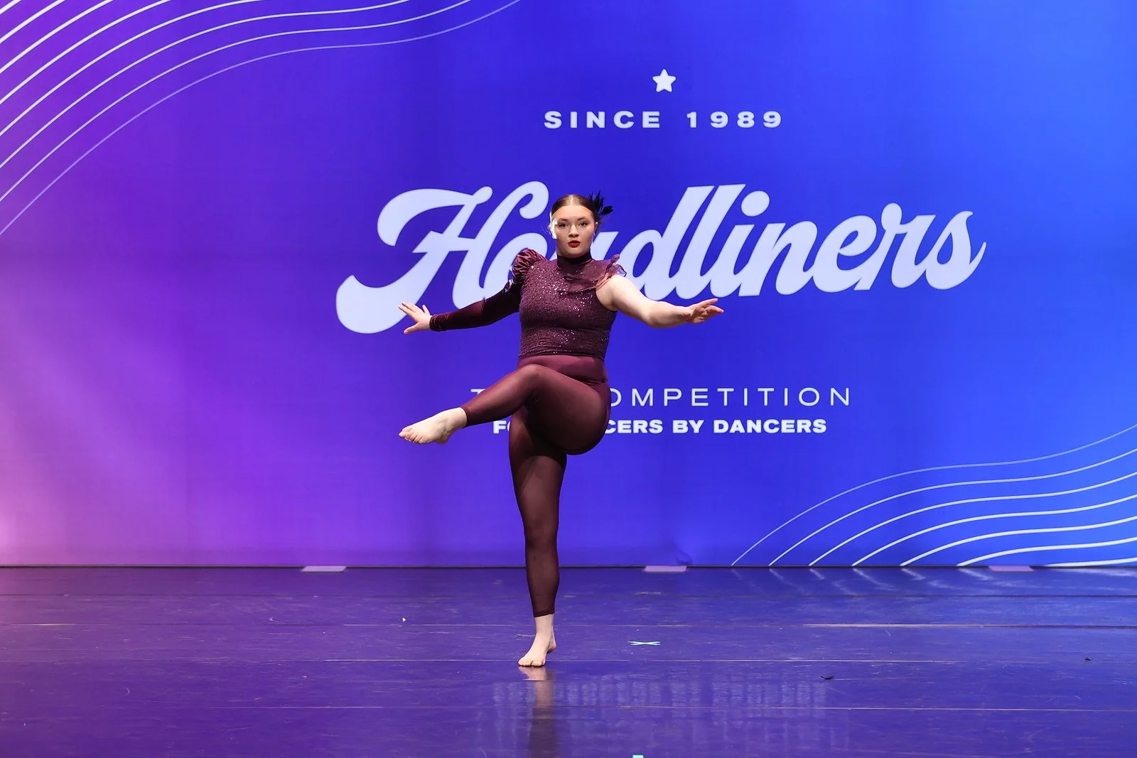 A dancer performing on stage at a competition called Holdinners, with a purple and blue background displaying the event's name and details.