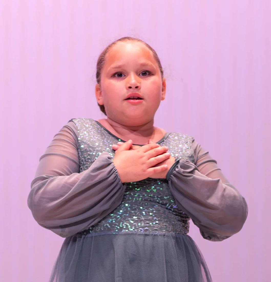 A young girl with her hands over her chest, wearing a gray dress with sequins, standing against a pinkish background.