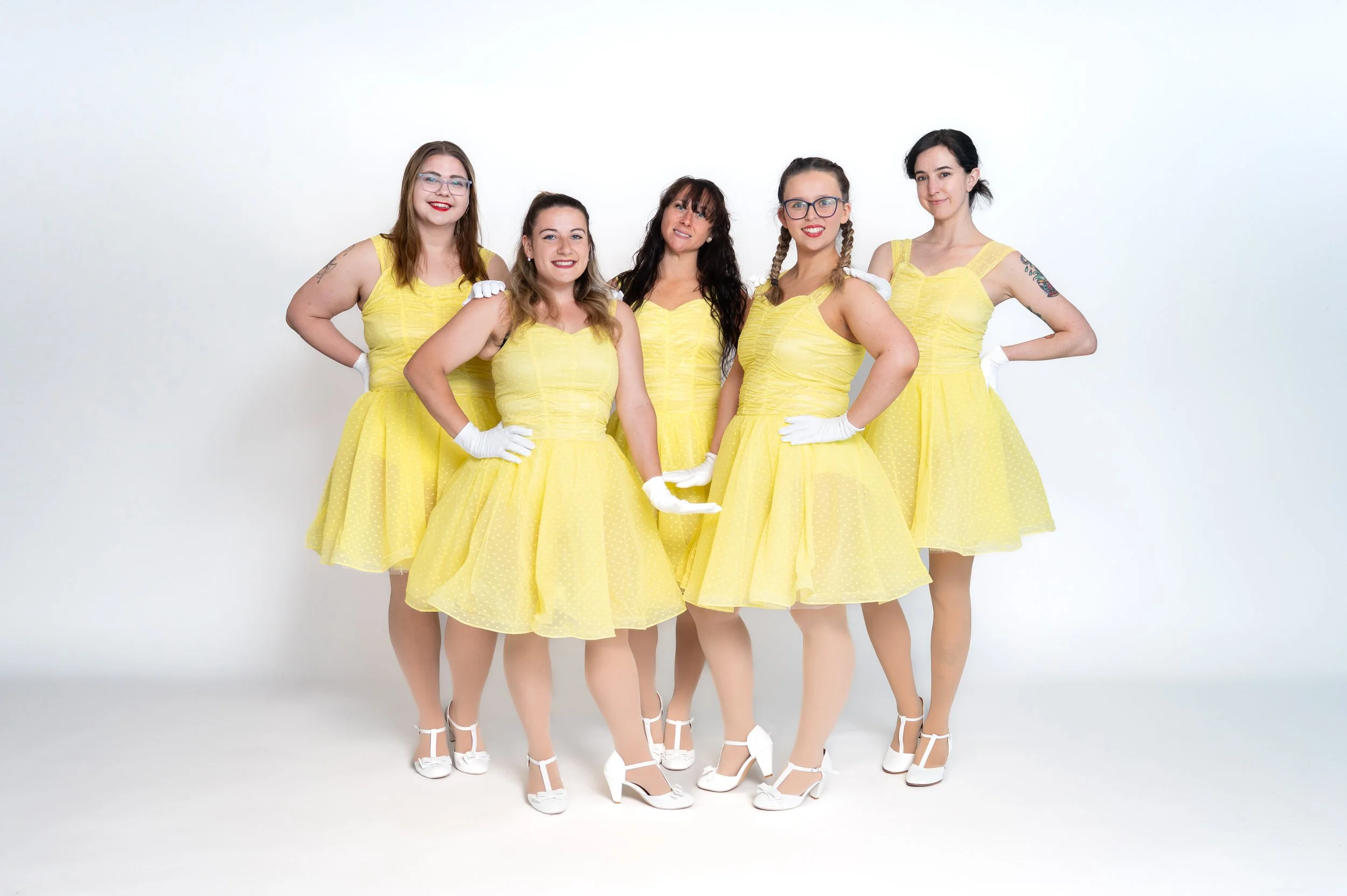 Five women in matching yellow vintage-style dresses and white heels, posing together in front of a plain white background.