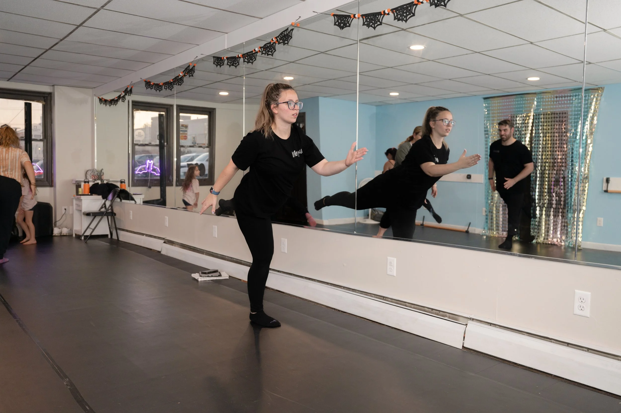A woman in black workout clothes balancing on one leg with arms outstretched in front of a mirror in a dance studio decorated with Halloween-themed garland. Other people and children are present in the studio.