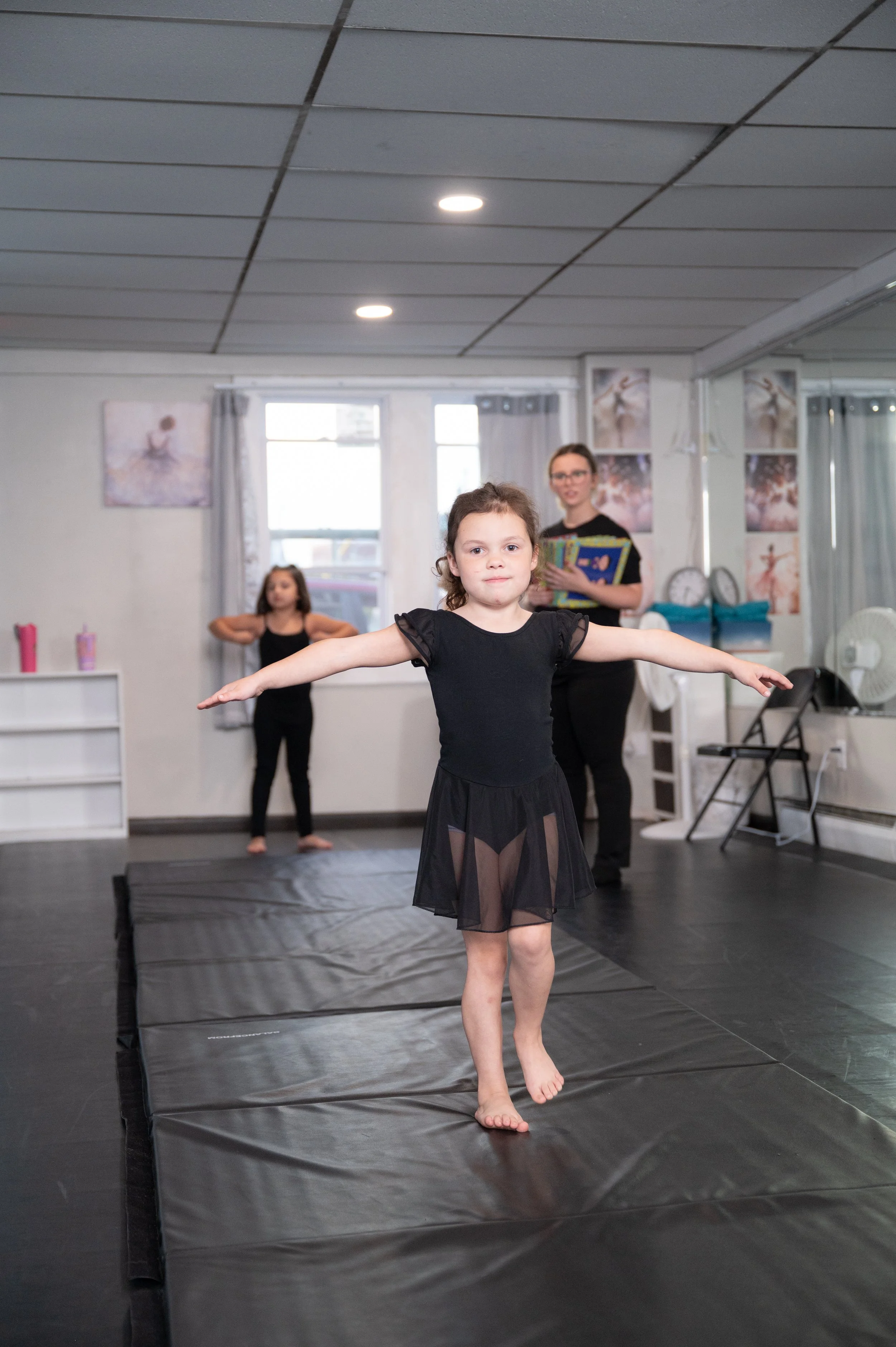 A young girl in a black dance costume practicing ballet on a mat in a dance studio, with an instructor and another girl in the background.