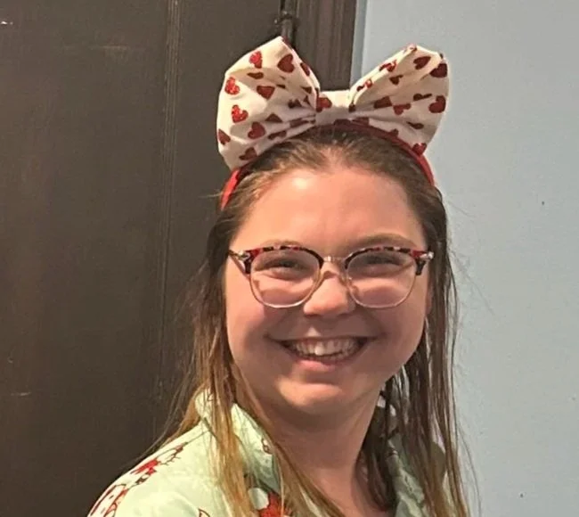 Woman smiling, wearing glasses and a headband with a bow patterned with hearts.