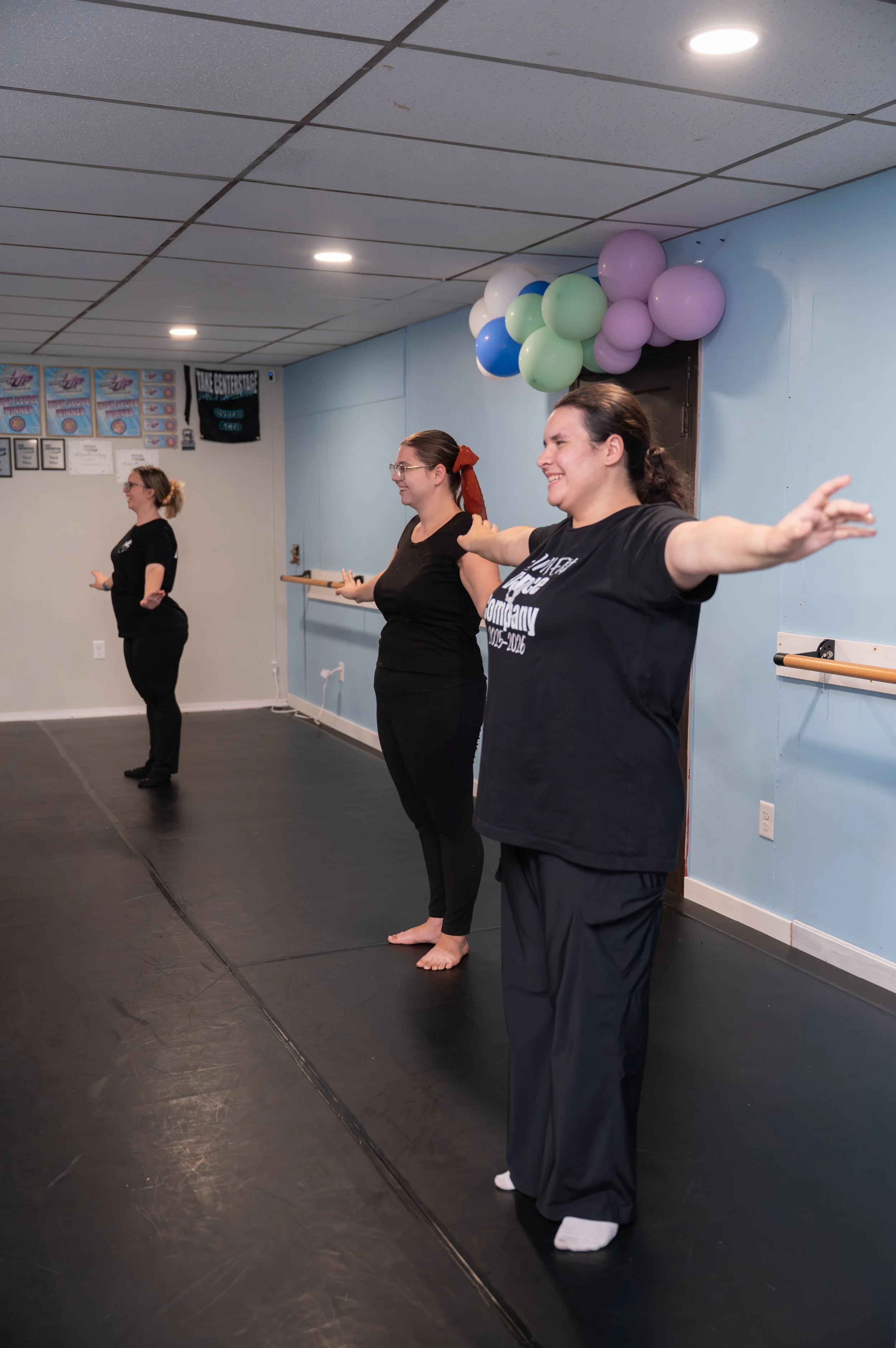 Three women stand in a dance studio with arms outstretched, smiling, facing forward. Decorated with balloons and posters on the wall.