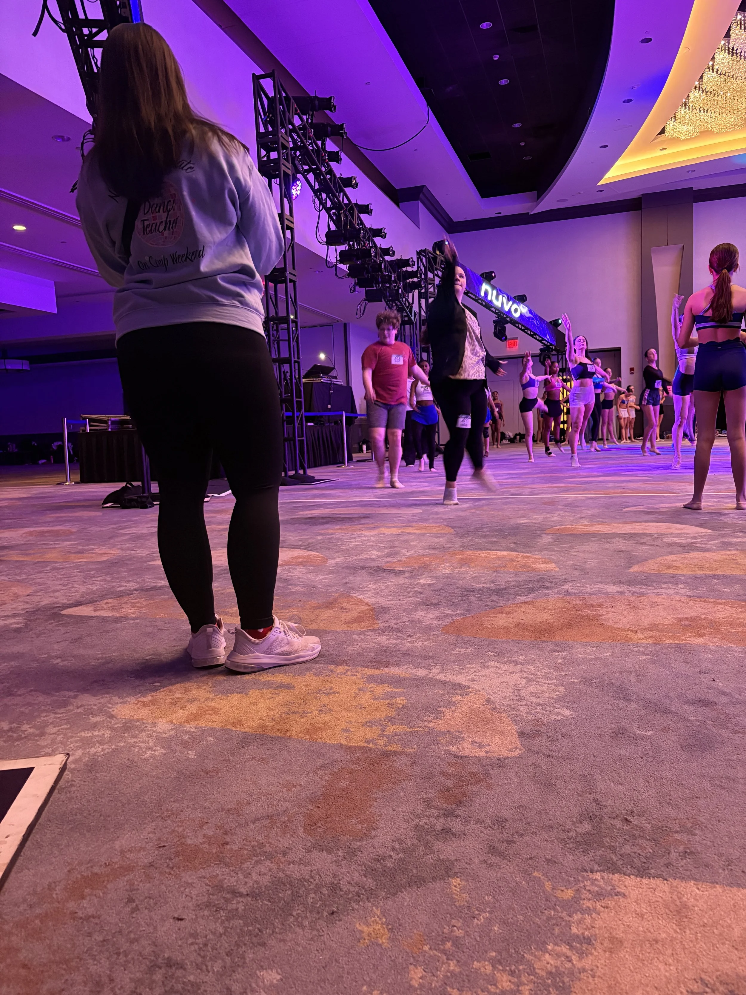 People participating in a dance or fitness class in a large, colorful conference room with stage lighting and a chandelier.