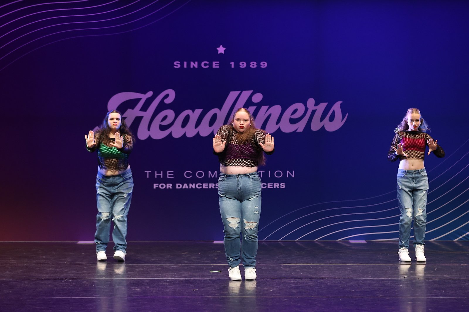 Three young female dancers on stage in casual attire, performing at the Headliners dance competition with a purple backdrop that reads 'Since 1989 Headliners, The Competition for Dancers.'