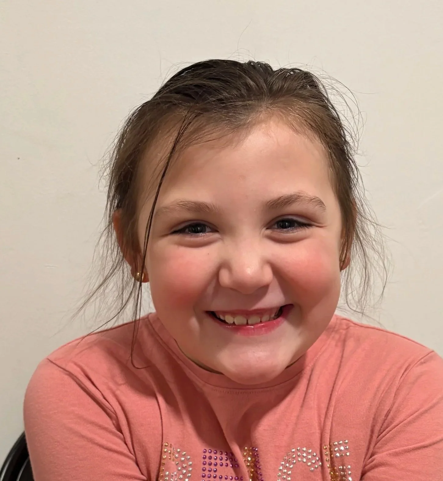 A young girl with brown hair, blue eyes, and a big smile, wearing a pink shirt, standing against a white background.