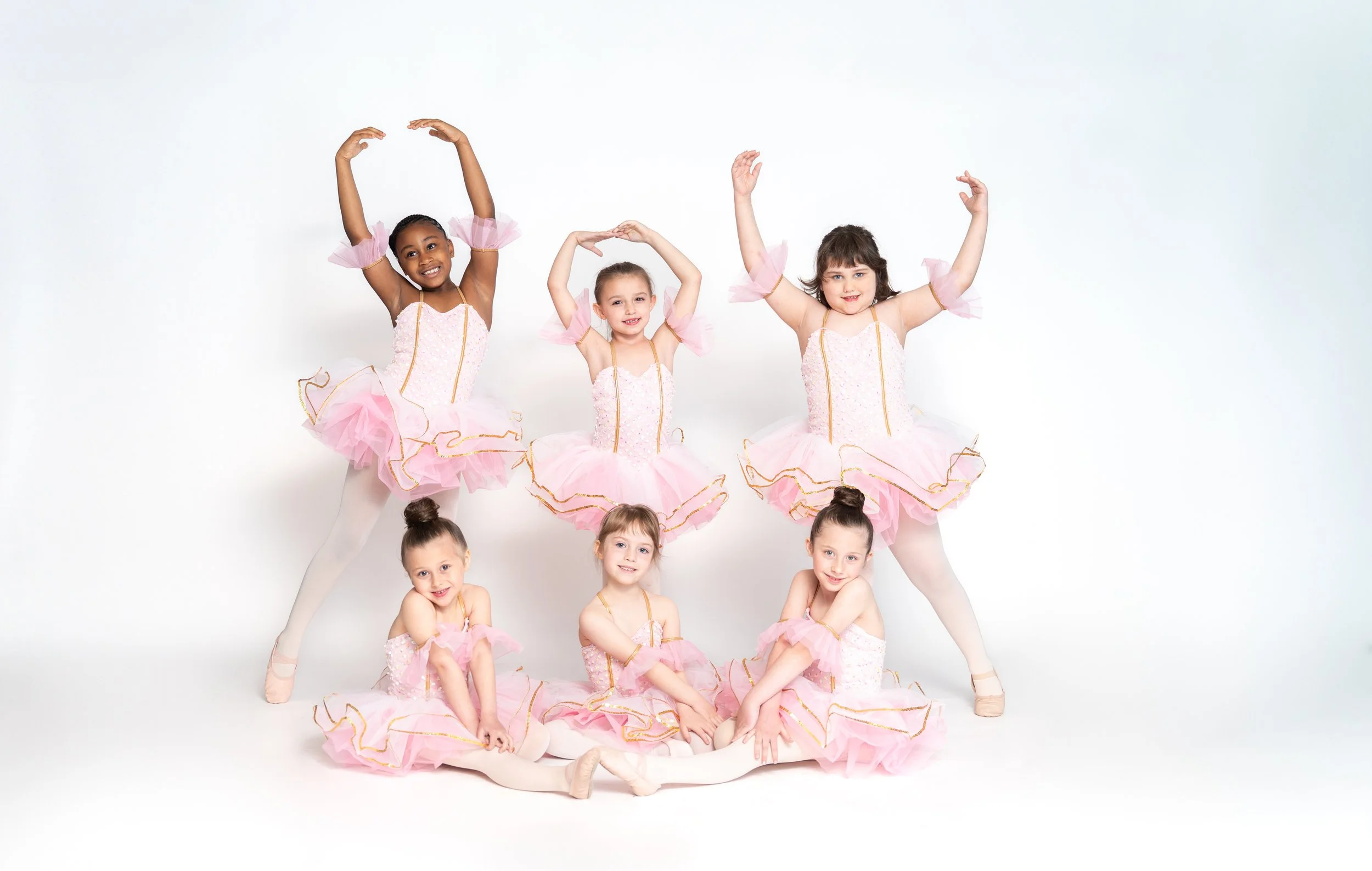 Seven young girls dressed in pink ballet tutus and leotards posing together in dance costumes against a white background, some sitting on the floor and others standing with arms raised.