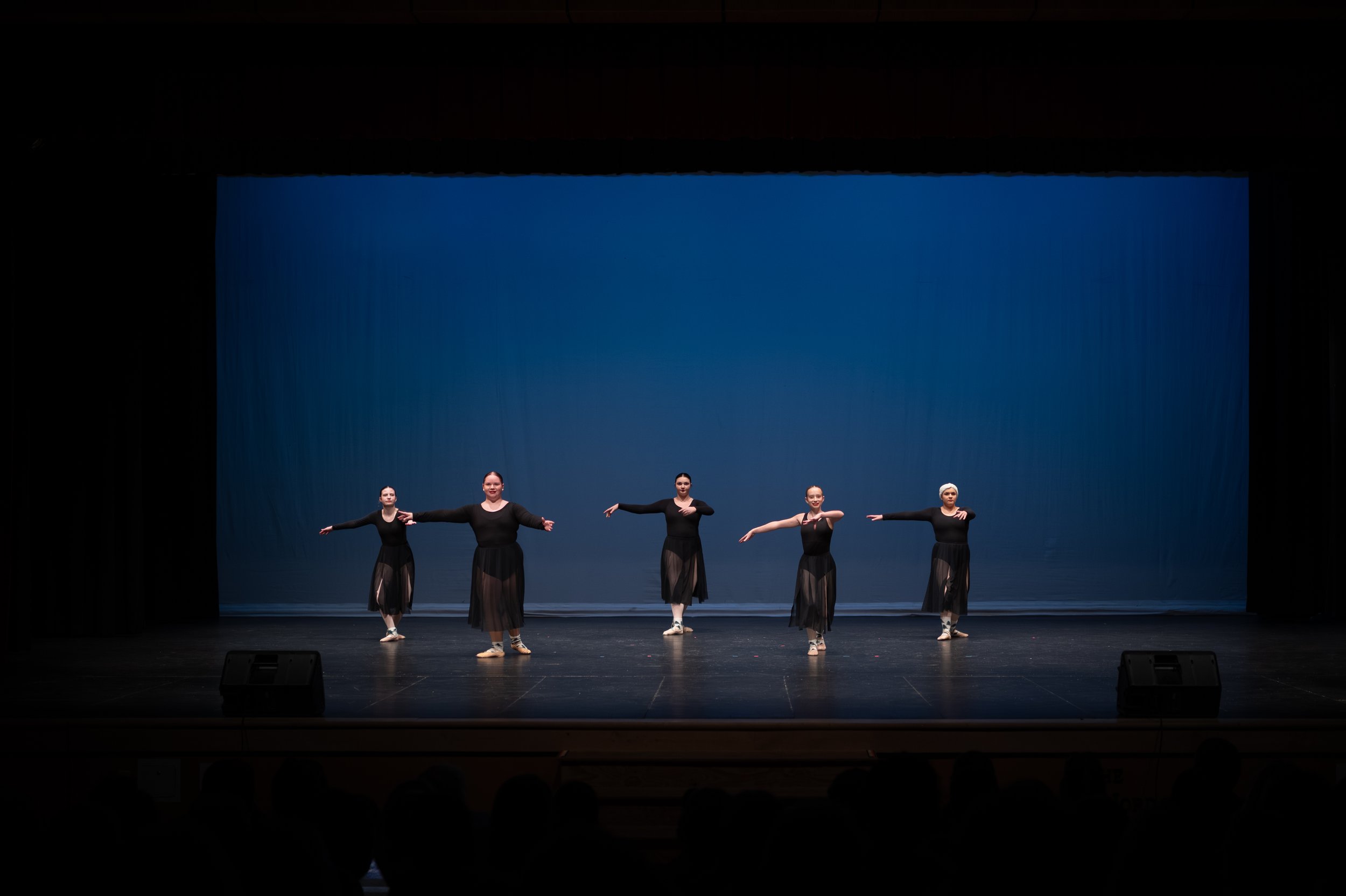 Five ballet dancers in black costumes perform on a stage with a blue backdrop.