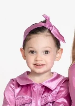 Close-up of a young girl with a pink satin bow headband and pink satin jacket, smiling at the camera.