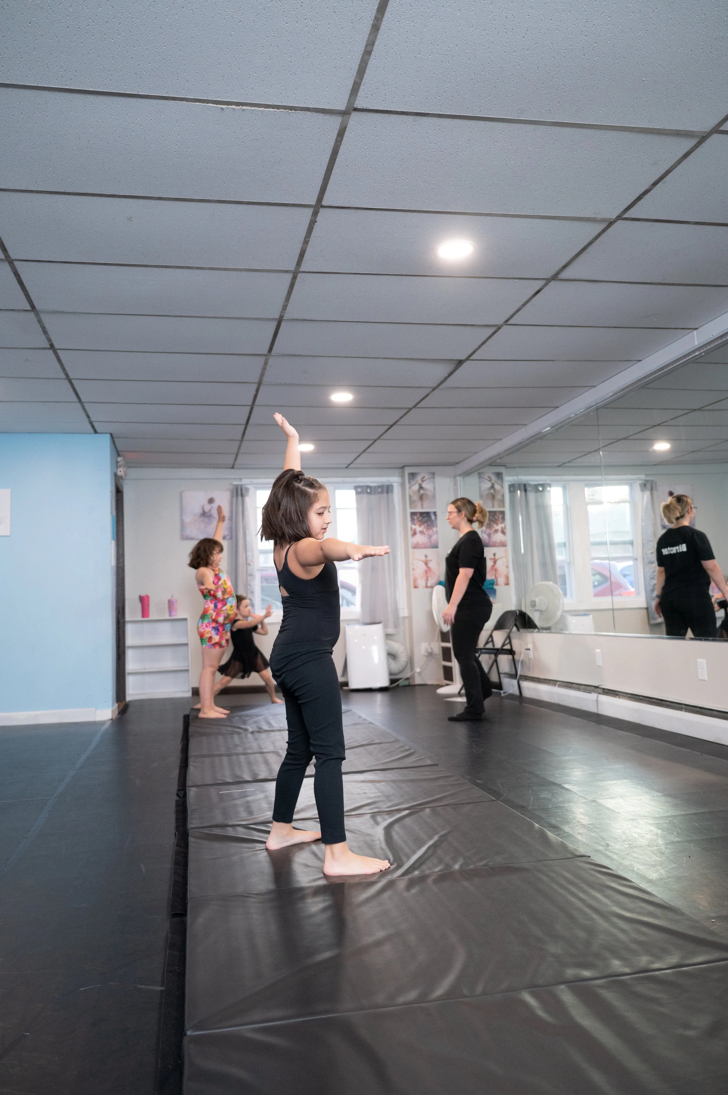 Children practicing ballet or dance moves on a padded floor in a dance studio, with an instructor overseeing them, mirrors on one wall, and posters on the walls.