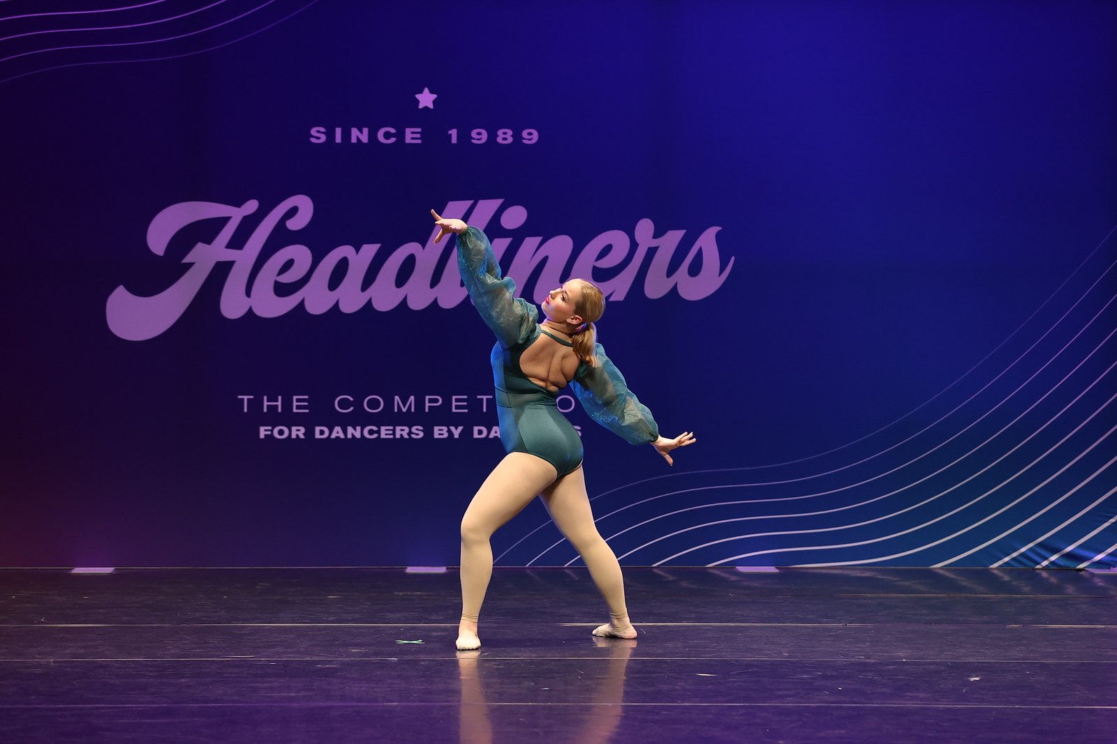 A dancer performing on stage with a dark purple backdrop that has the words 'Since 1989 Headliners' and 'The competition for dancers by dancers' printed on it. The dancer is in a mid-spin pose, wearing a teal and black costume with sheer sleeves and beige tights.