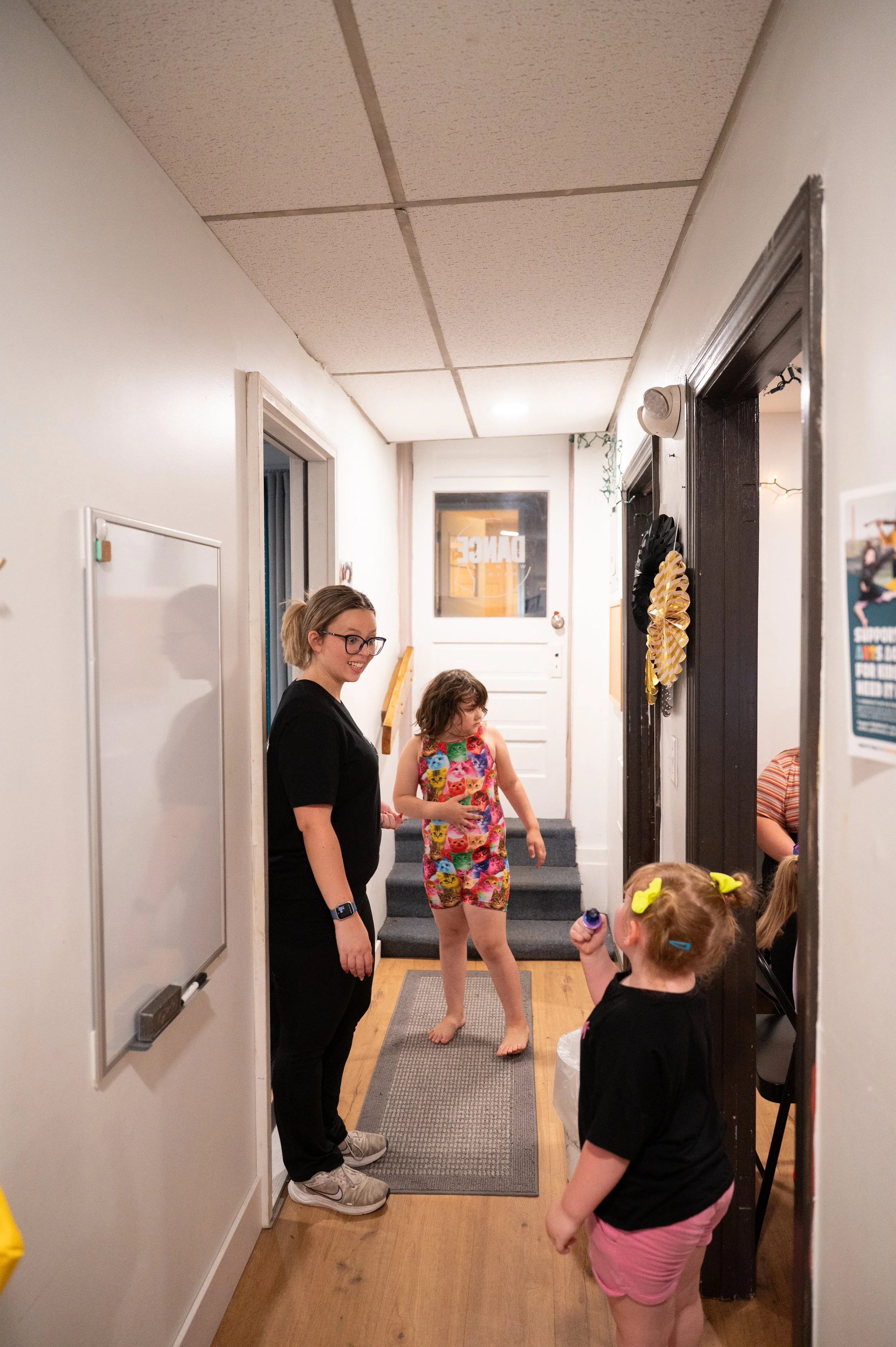 Children and woman standing in a hallway near a staircase and a door with an 'OPEN' sign. One girl with yellow bows in her hair holding a bottle, another girl in floral dress, woman wearing black.