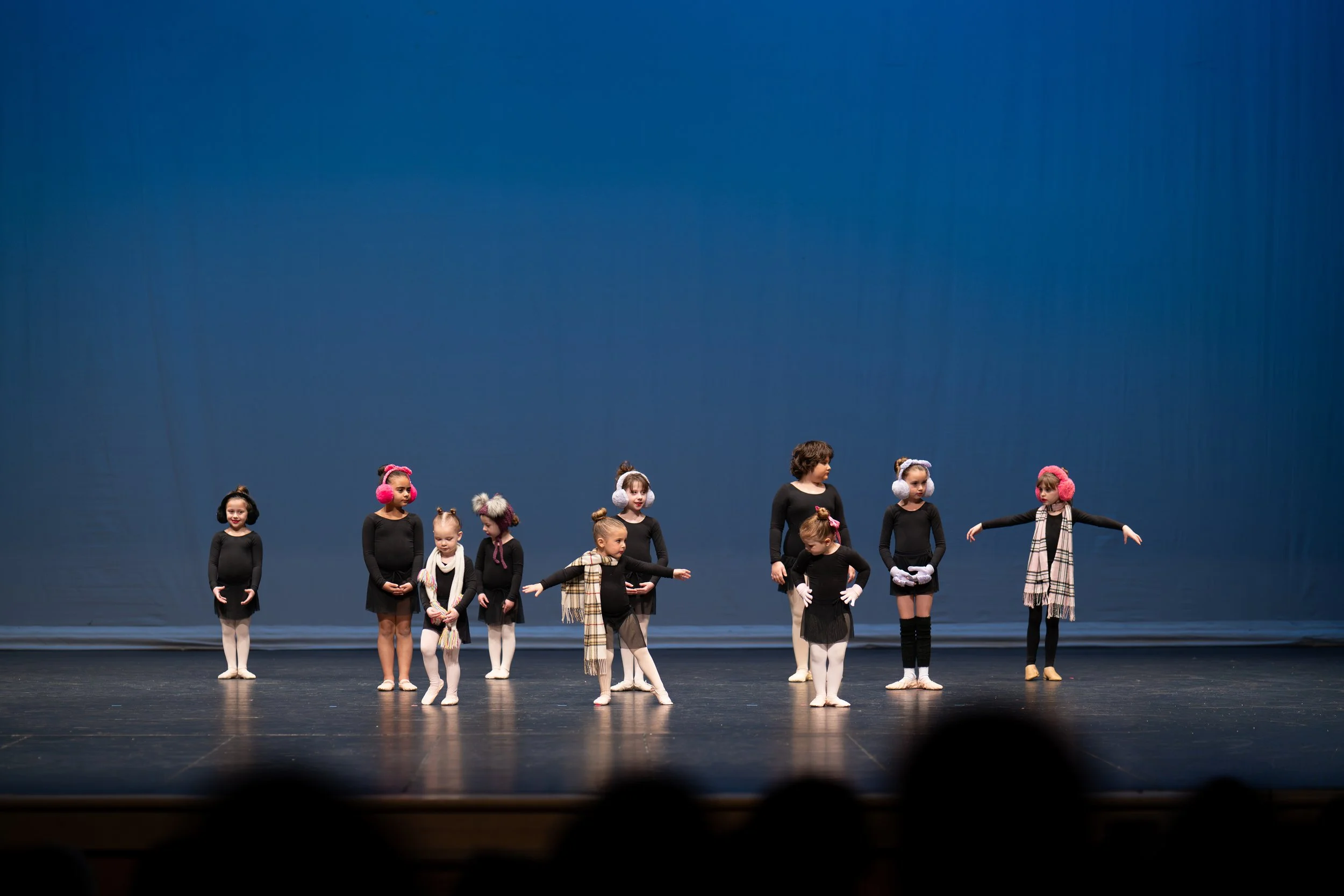Children performing a ballet on stage, wearing black leotards, tights, and earmuffs, with some also wearing scarves and gloves, against a plain blue background.