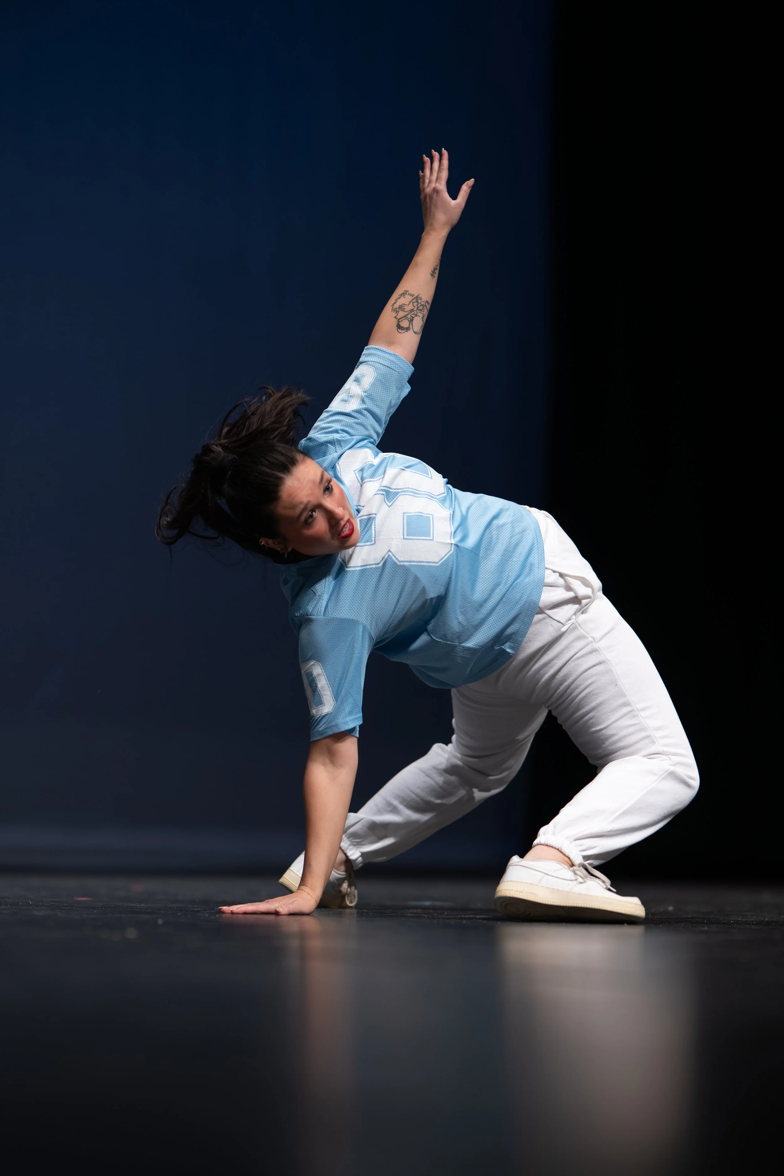 A woman in a blue sports jersey and white pants in a dance pose on stage, with one hand on the floor and the other raised above her head, looking towards the camera.