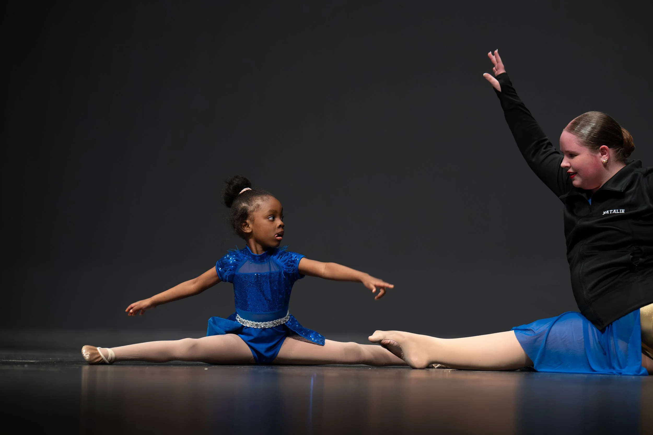A young girl in a blue dance costume doing a split on stage, reaching out towards an instructor who is stretching her leg in a split, both practicing ballet or gymnastics.