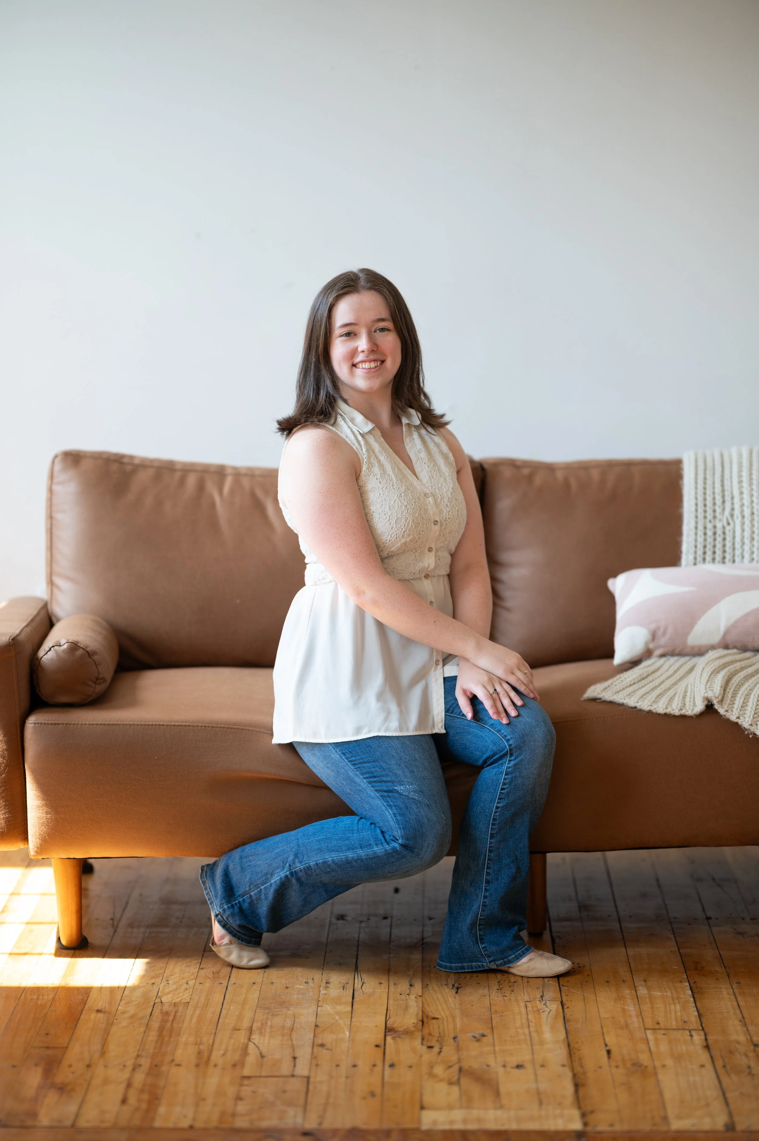 A woman with shoulder-length dark hair, wearing a sleeveless cream blouse and blue jeans, sitting on a brown sofa in a sunlit room, smiling at the camera.