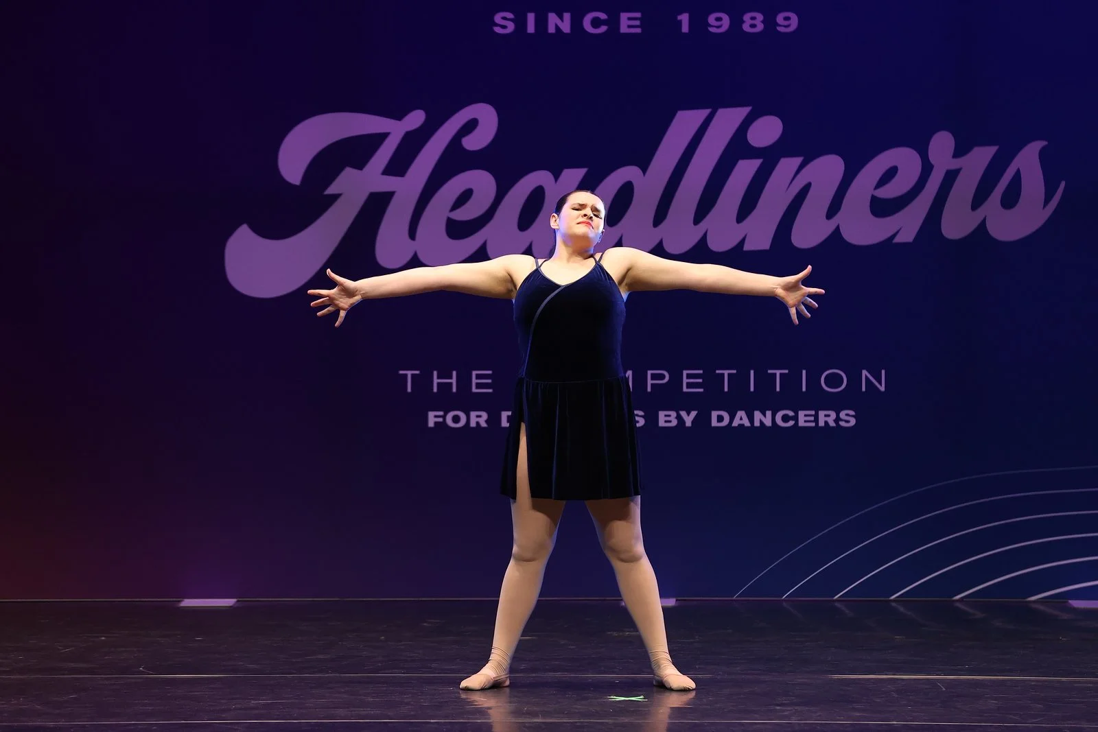 A ballet dancer standing on stage with arms outstretched, wearing a dark dress, during a dance competition.