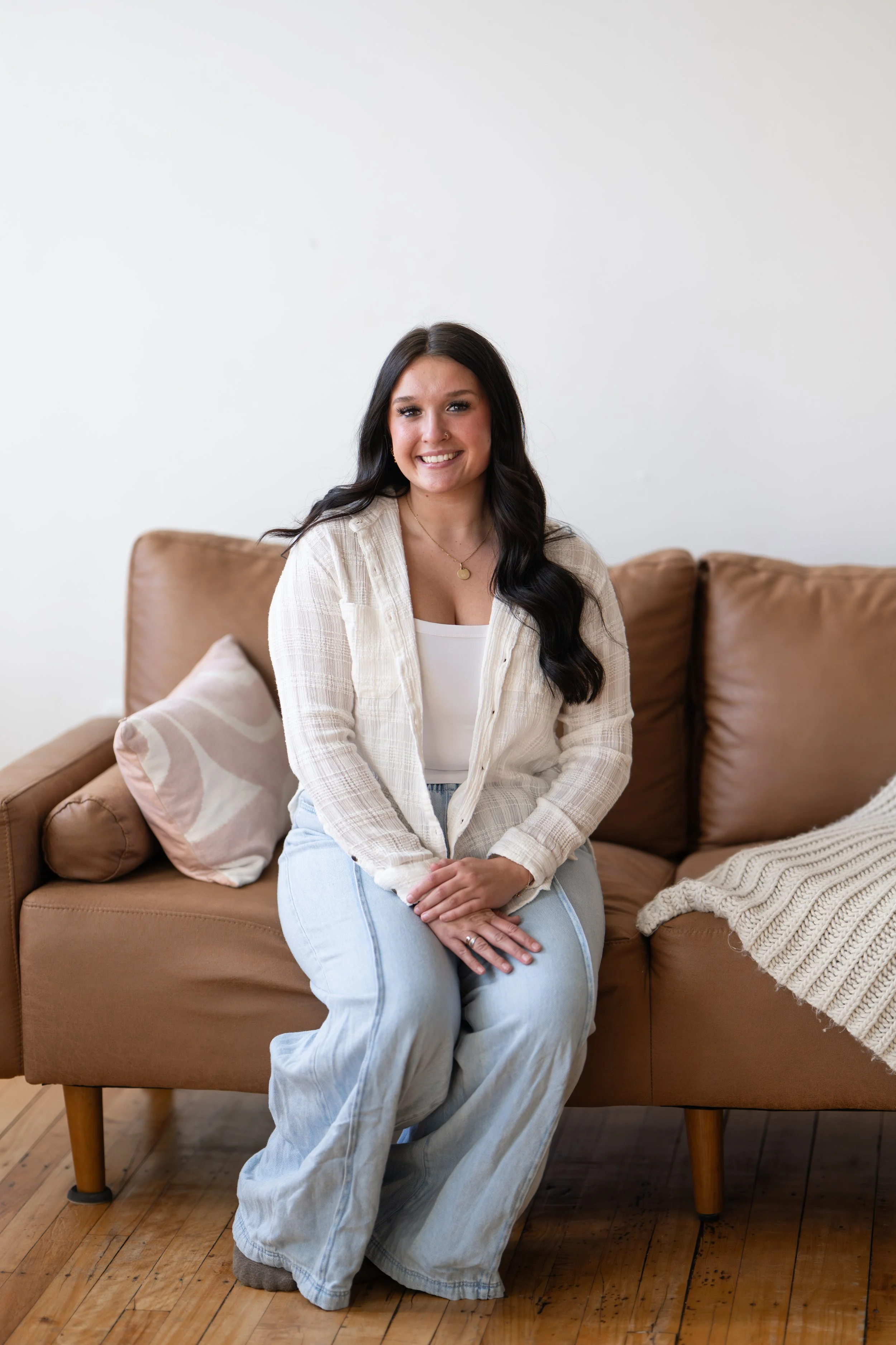 A woman with long dark hair, wearing a white shirt and a beige checked shirt, sitting on a beige sofa with a pillow and a blanket, smiling in a bright room with wooden floor.