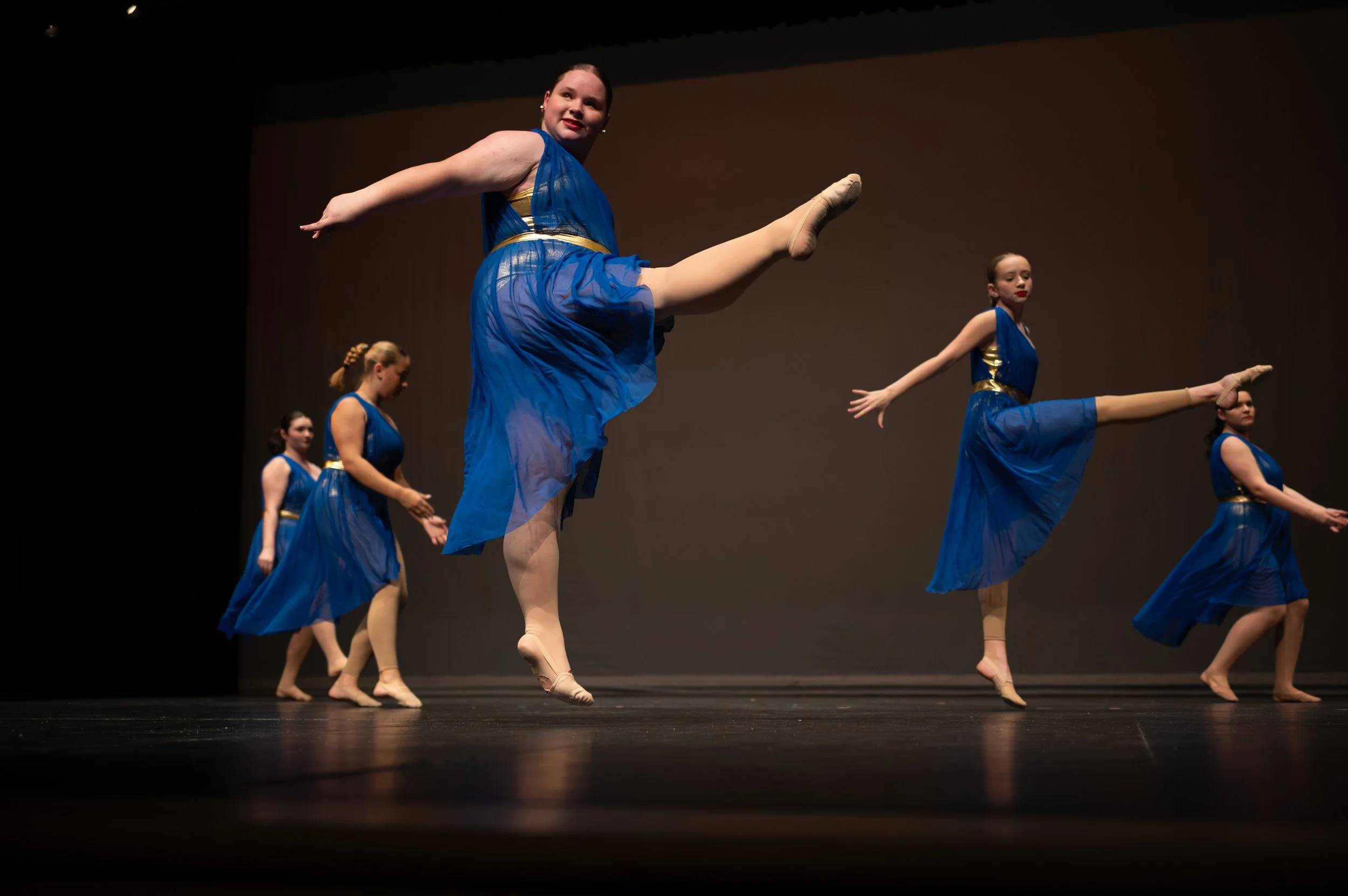 Five young girls in blue costumes performing a ballet dance on stage. They are mid-movement, with some jumping and others standing, against a dark background.