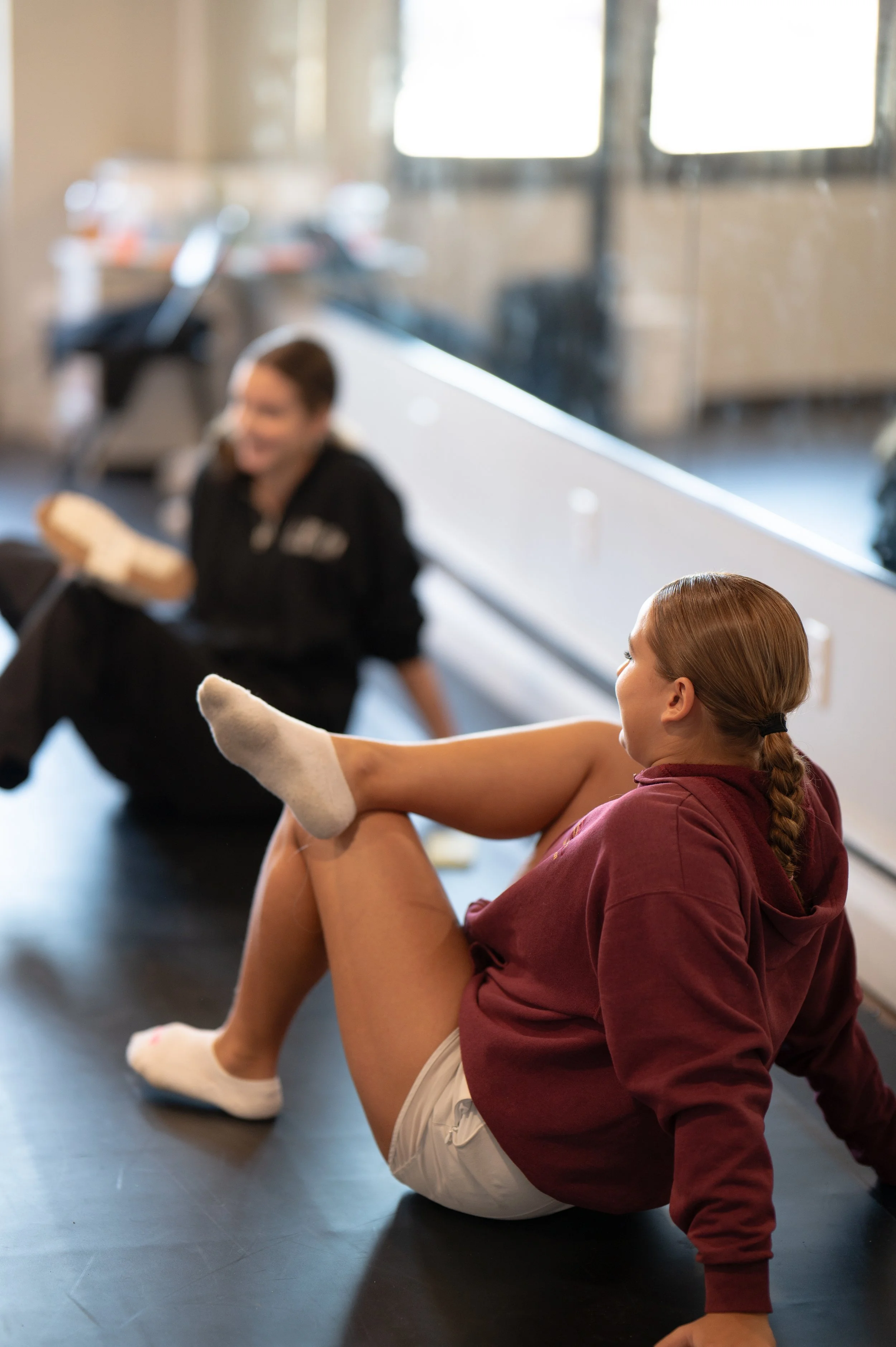 A young woman in athletic clothes and socks sitting on the floor of a gym, stretching her leg while a trainer in black clothing sits nearby holding a water bottle and a towel.