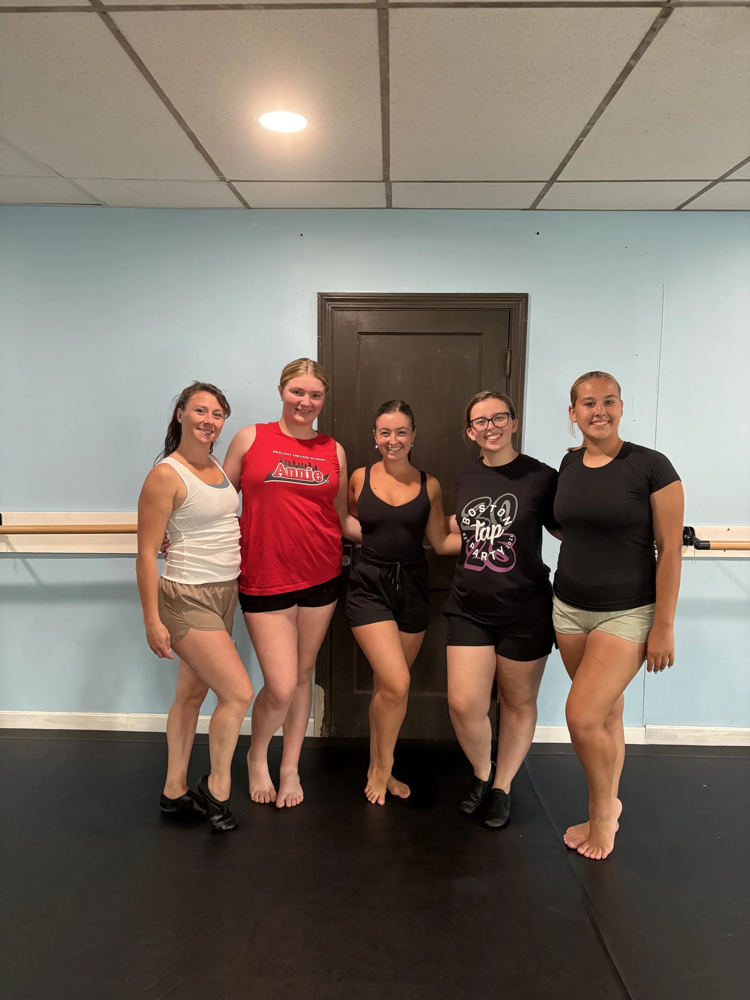 Group of five women standing in a dance studio, smiling at the camera, some with arms around each other.