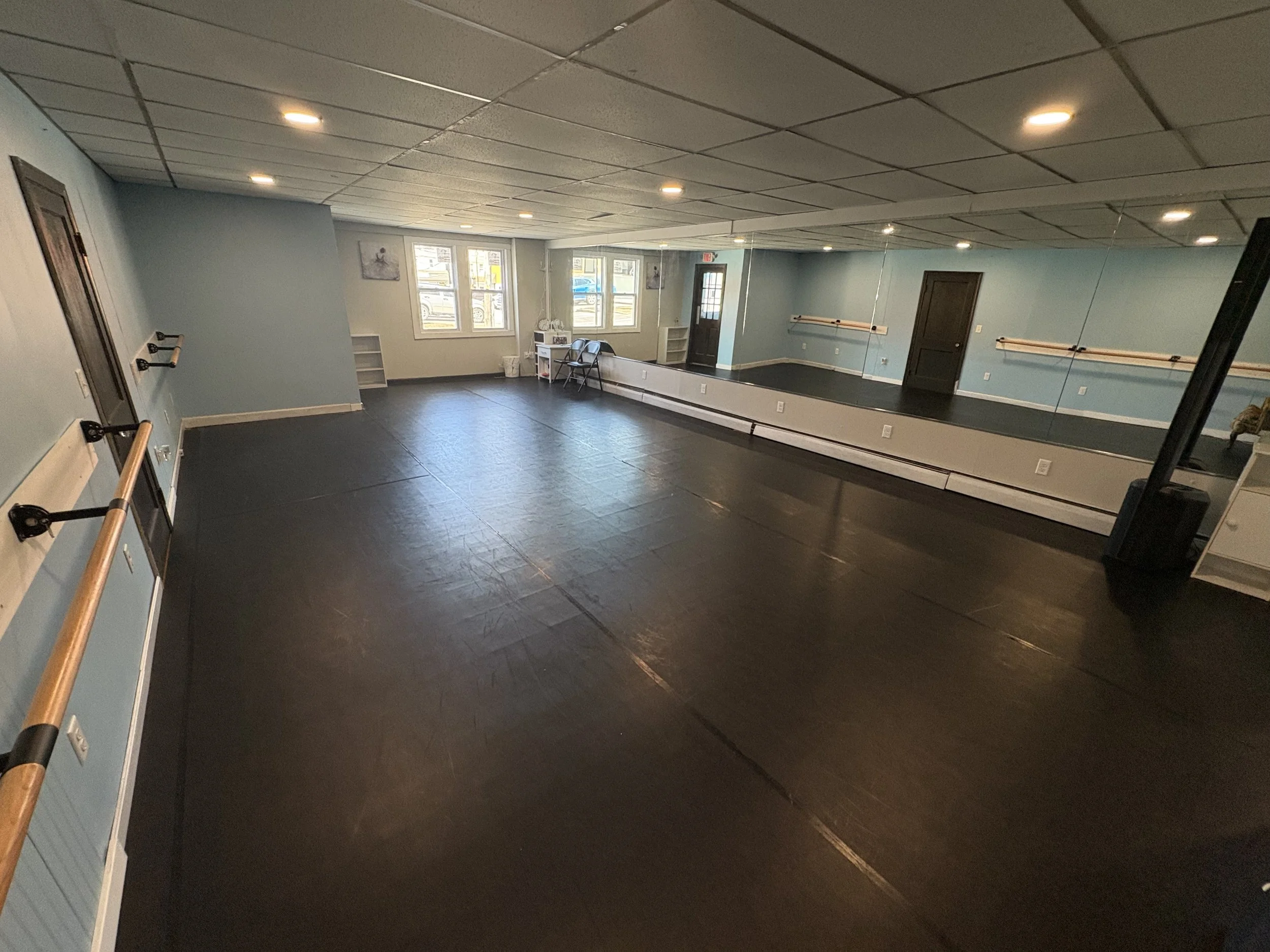 A spacious dance or exercise studio with black flooring, light blue walls, and ceiling with recessed lighting. Large mirror spans one wall, with ballet barres along it. Windows let in natural light. A door and some chairs are visible near the windows.