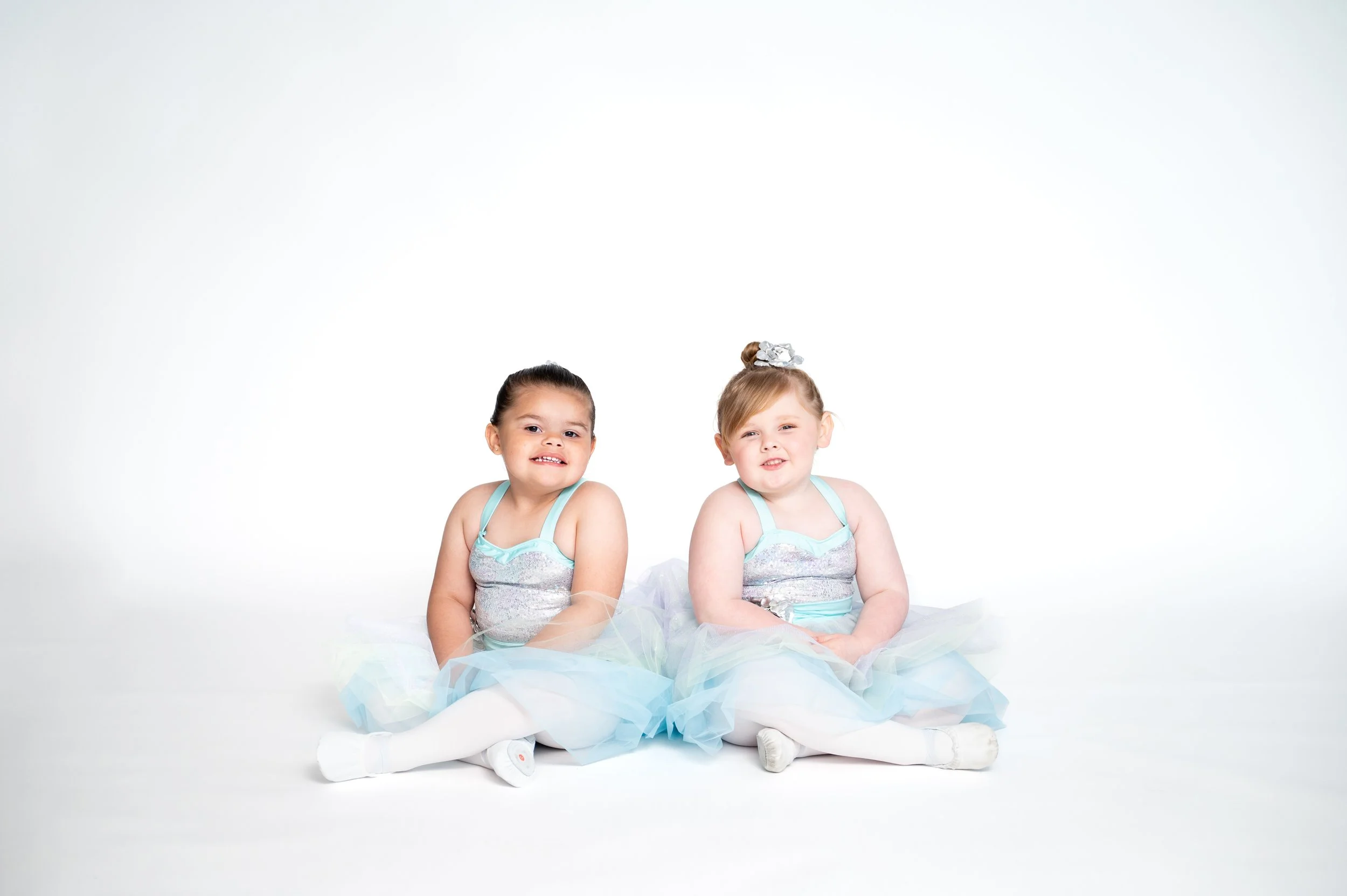Two young girls in ballet costumes sitting on the floor against a white background.