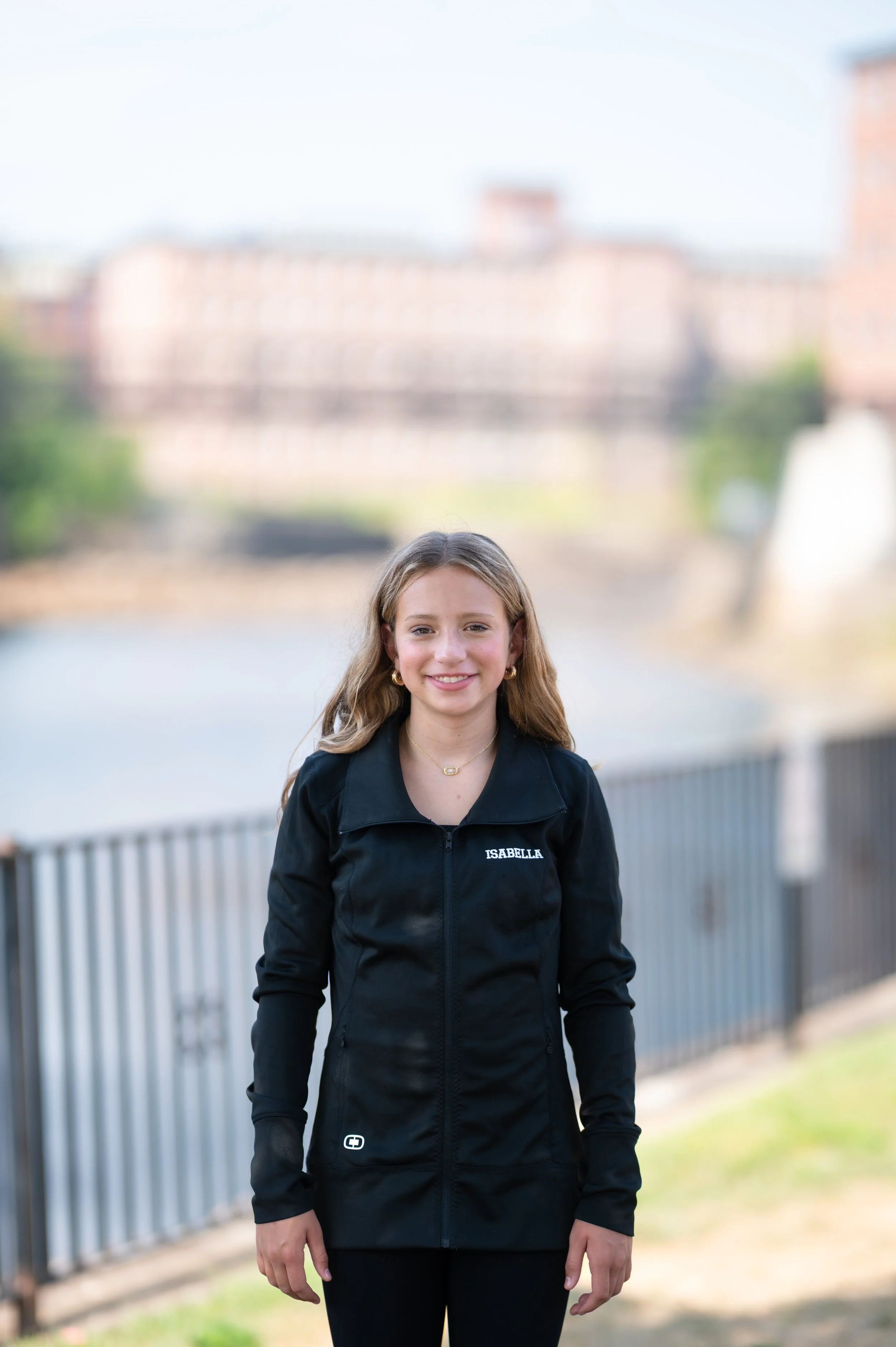 A young girl with long, wavy blonde hair smiling outdoors near a river, wearing a black jacket with her name 'Isabella' embroidered on it.