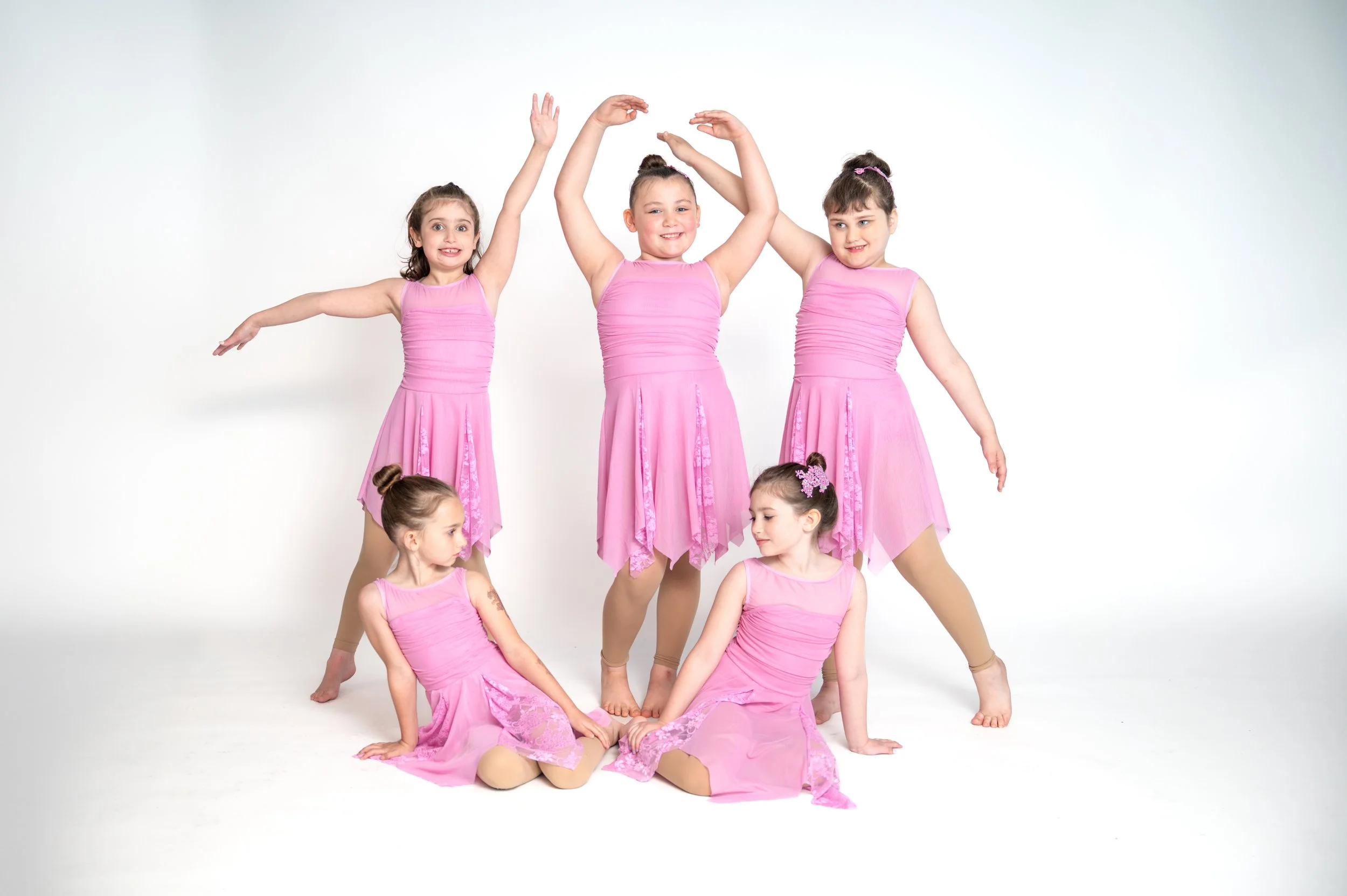 Six young girls dressed in pink ballet costumes posing together against a white background, with some standing and others sitting on the floor, engaging in a dance or ballet performance.
