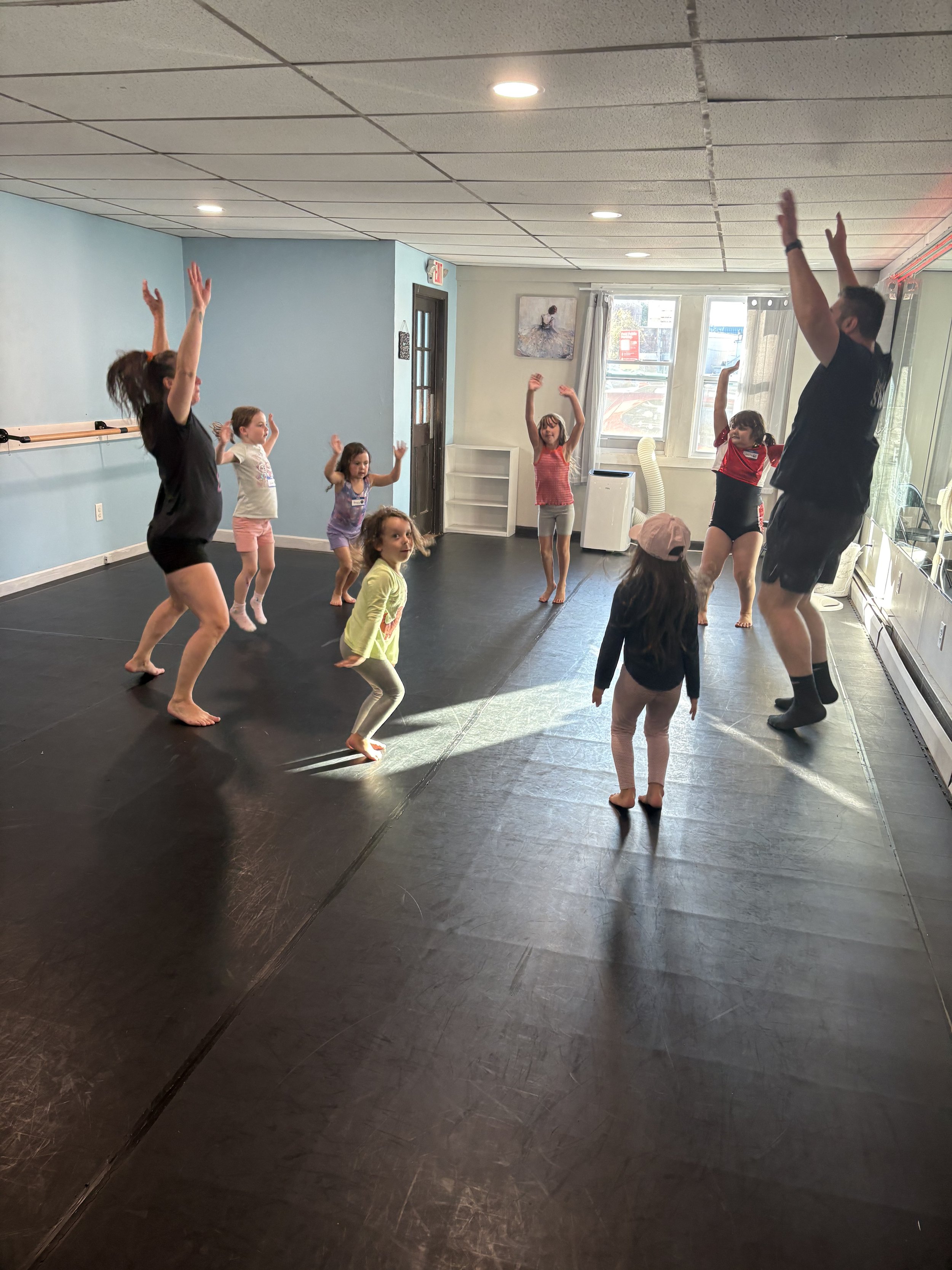 Children and instructor participating in an indoor dance or exercise class, with raised arms, on a black sprung floor, in a room with blue and white walls and large windows letting in natural light.