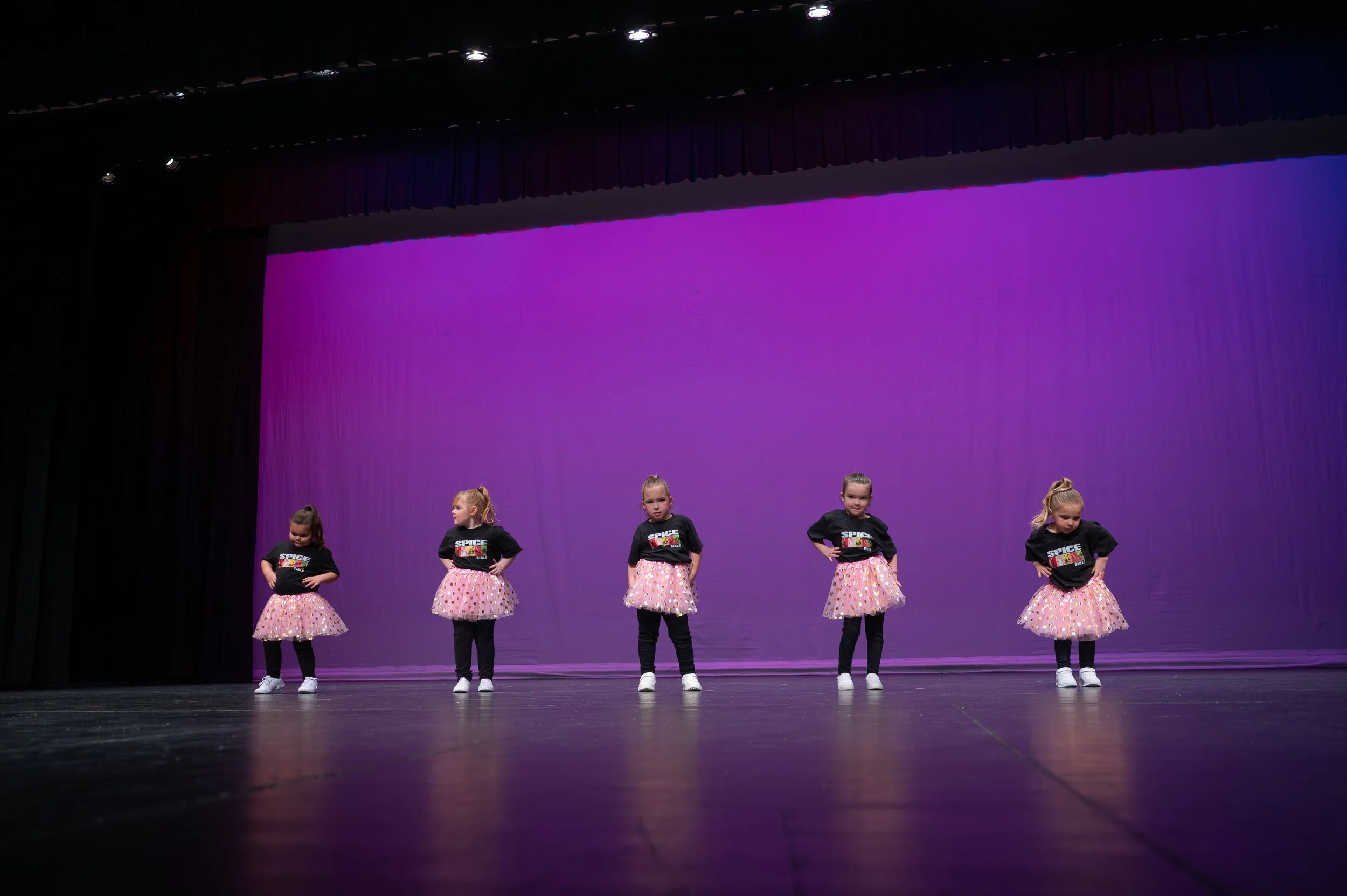 Five young girls performing a dance on stage with purple backdrop, wearing matching black T-shirts with a graphic and pink polka dot tutus.