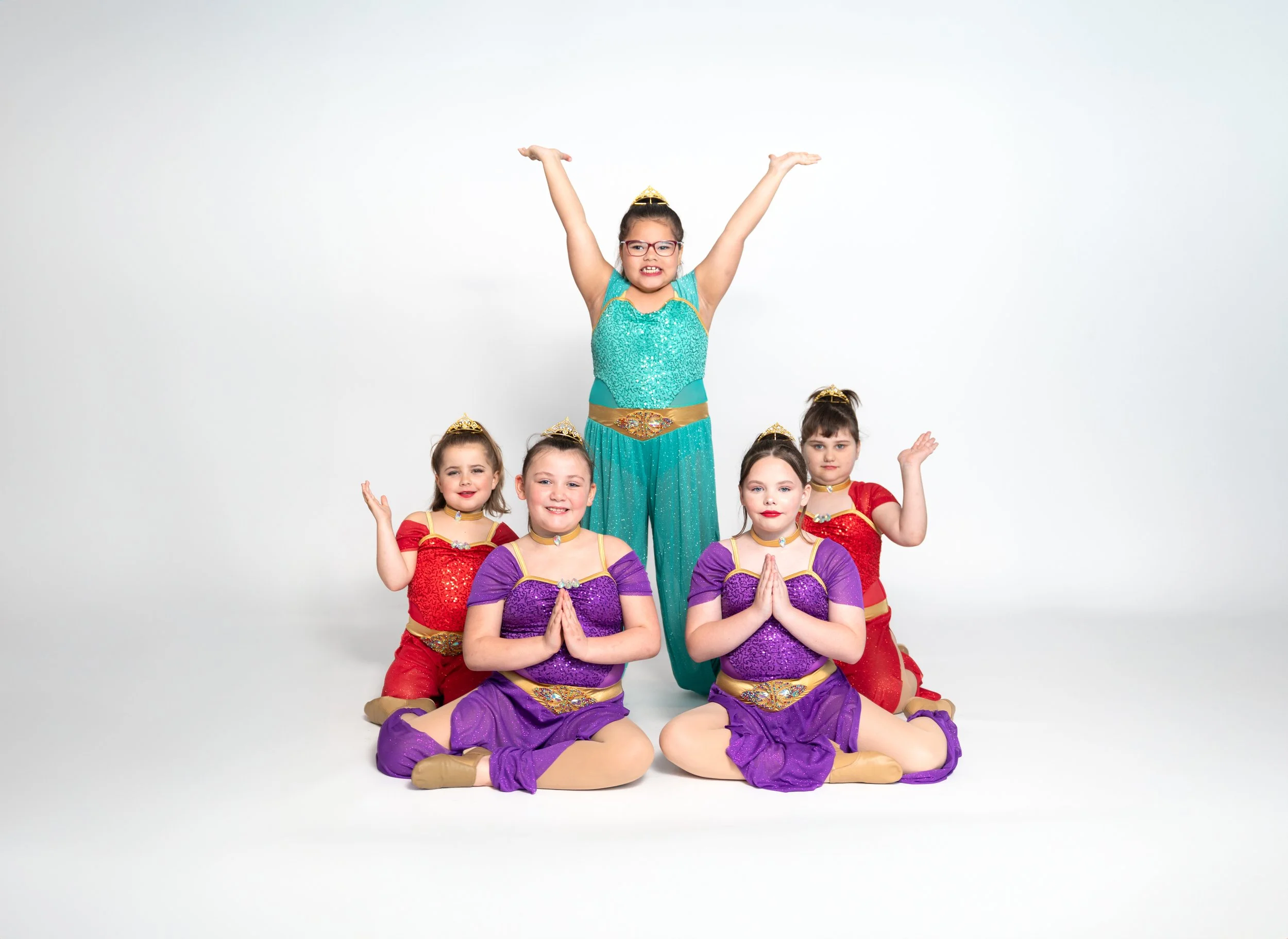 Five young girls dressed in colorful costumes, some sitting cross-legged and others kneeling, posing with hands in prayer position, against a plain white background.