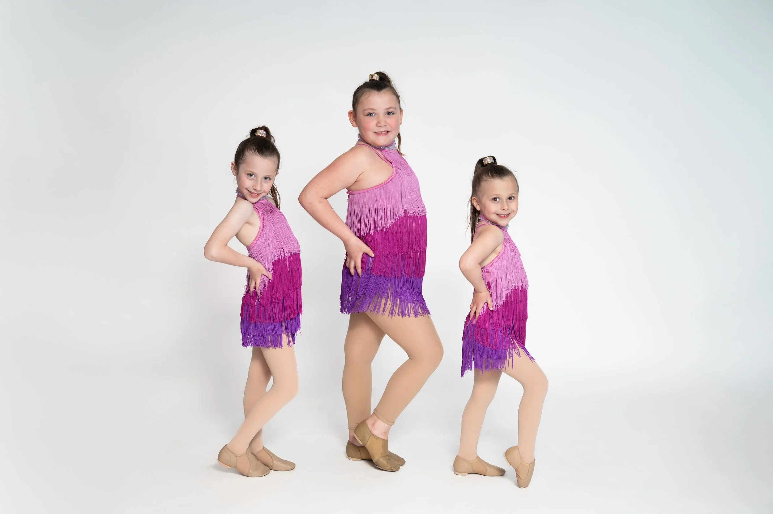 Three young girls dressed in matching pink and purple fringed dance costumes pose with hands on hips against a white background.