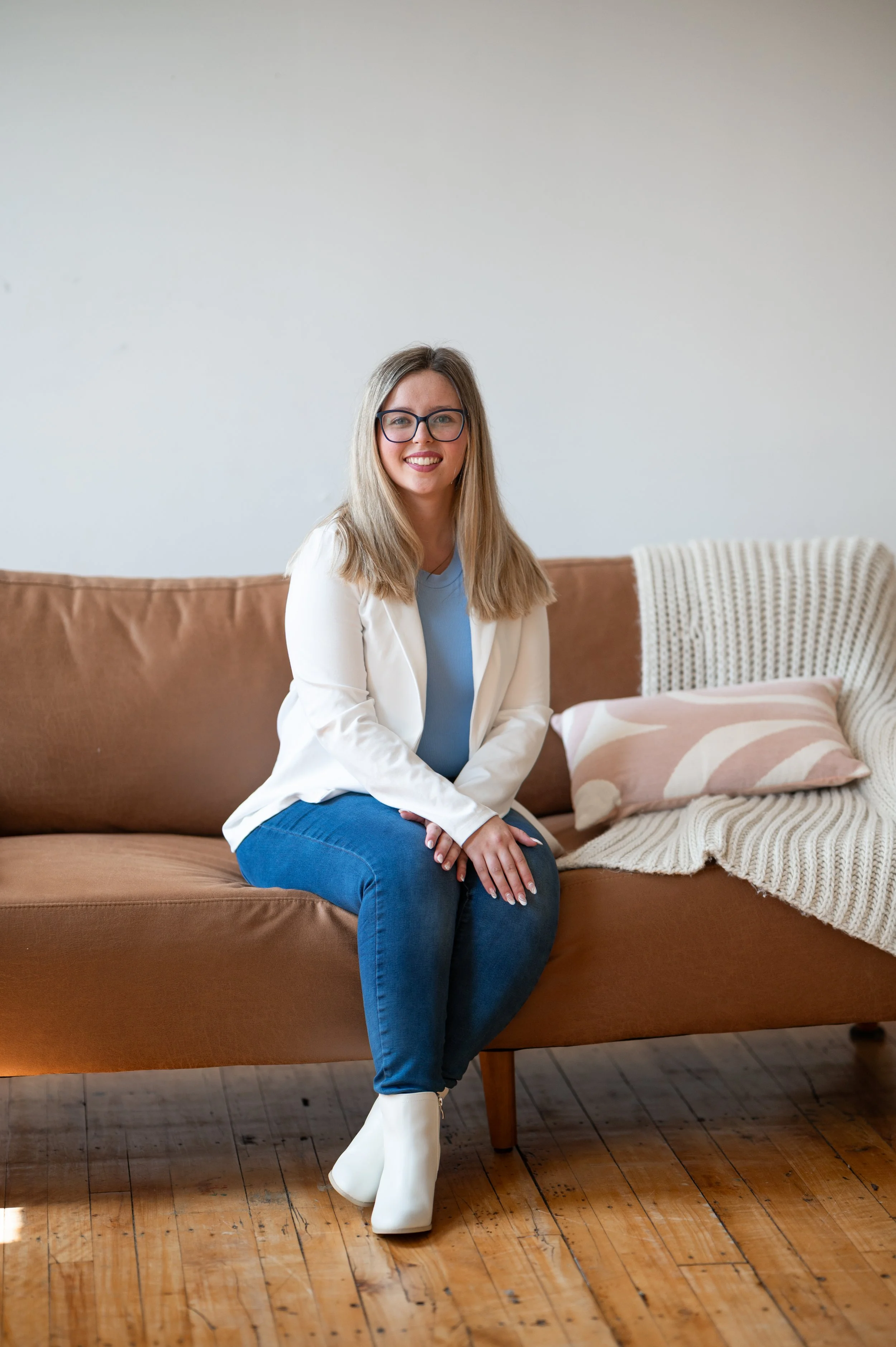 A woman with blonde hair and glasses sitting on a brown sofa, smiling, in a room with wooden flooring and a white wall. She is wearing a white blazer, blue top, blue jeans, and white boots.