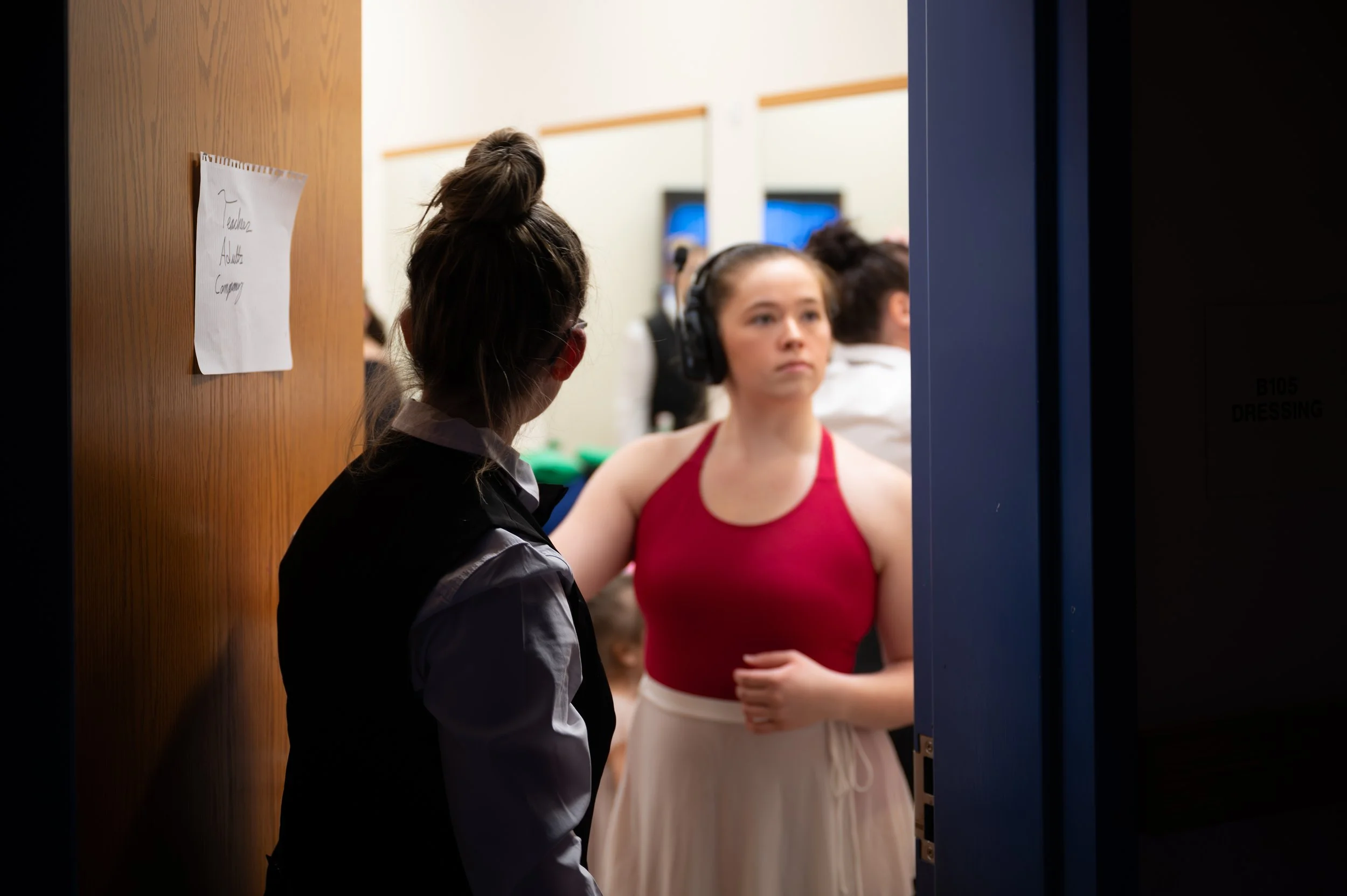 A young woman in a red top and white skirt standing in a rehearsal room, facing a person with their back turned, near a blue door, with other people blurred in the background.