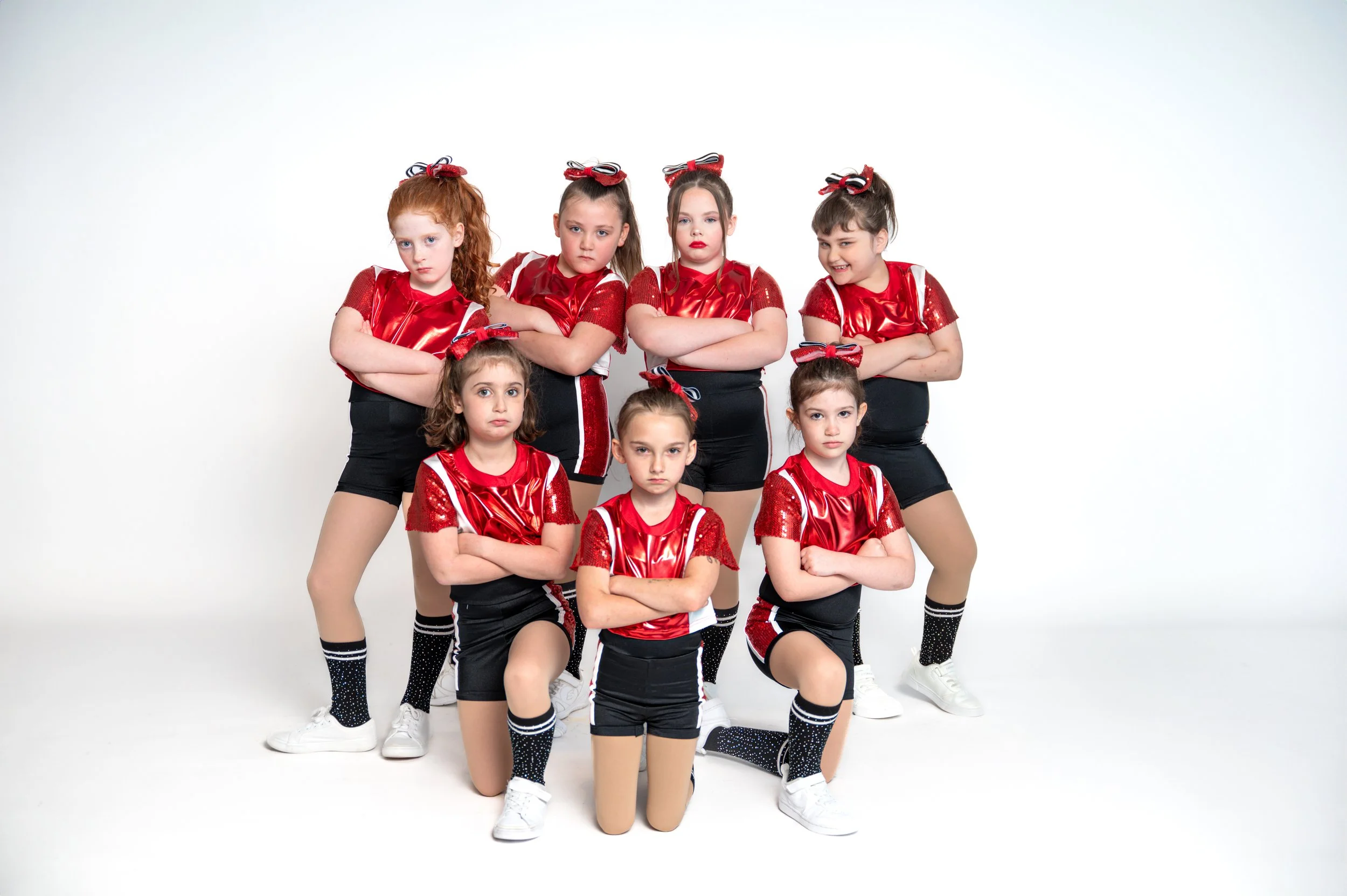 A group of seven young girls in red and black cheerleading uniforms with bows in their hair, posing with folded arms against a white background.