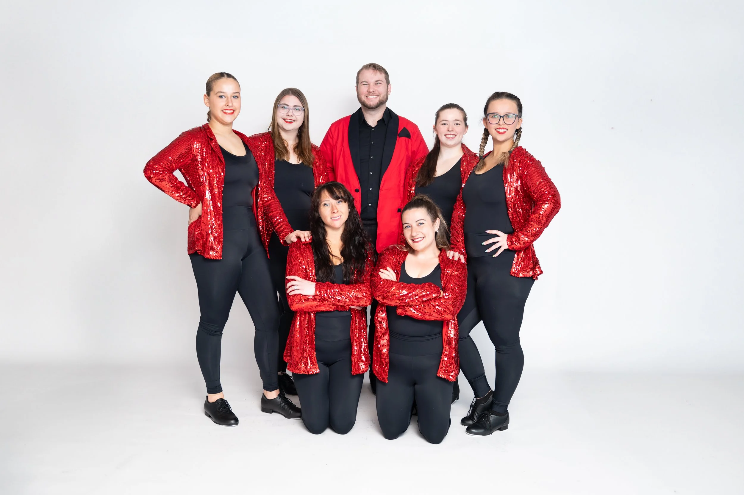 Group of seven performers in coordinated red and black costumes, smiling and posing against a white background.