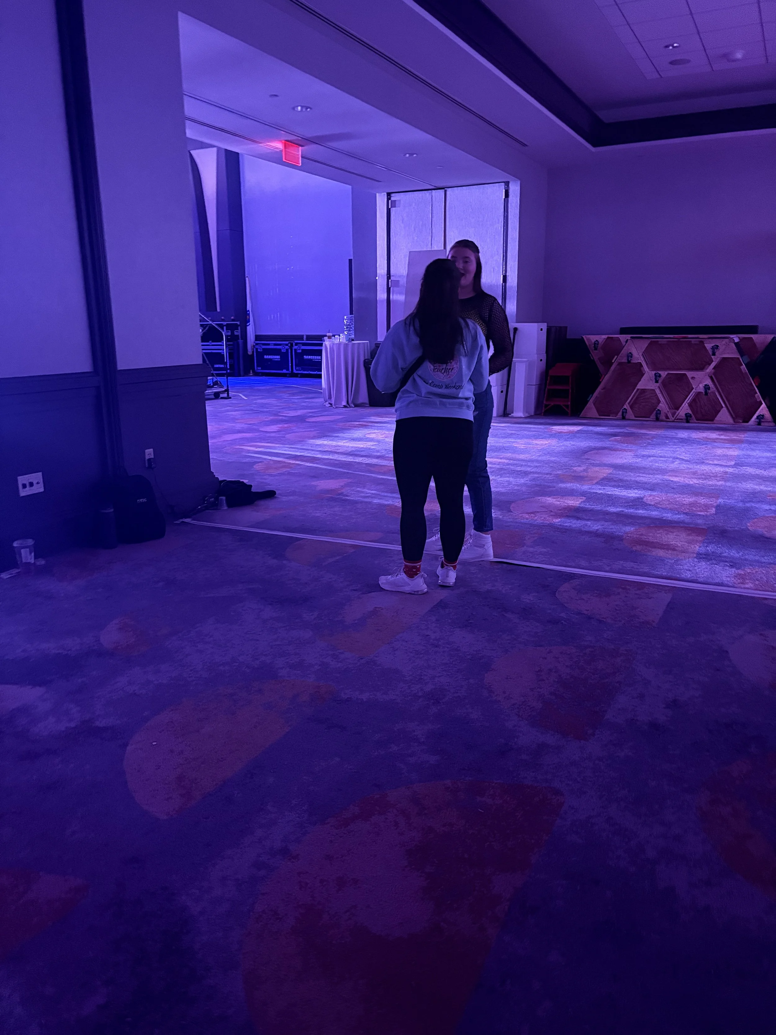 Two women talking at an event in a large, dimly lit room with purple lighting. One woman is wearing a gray hoodie and black pants, and the other woman is wearing a black top and jeans.