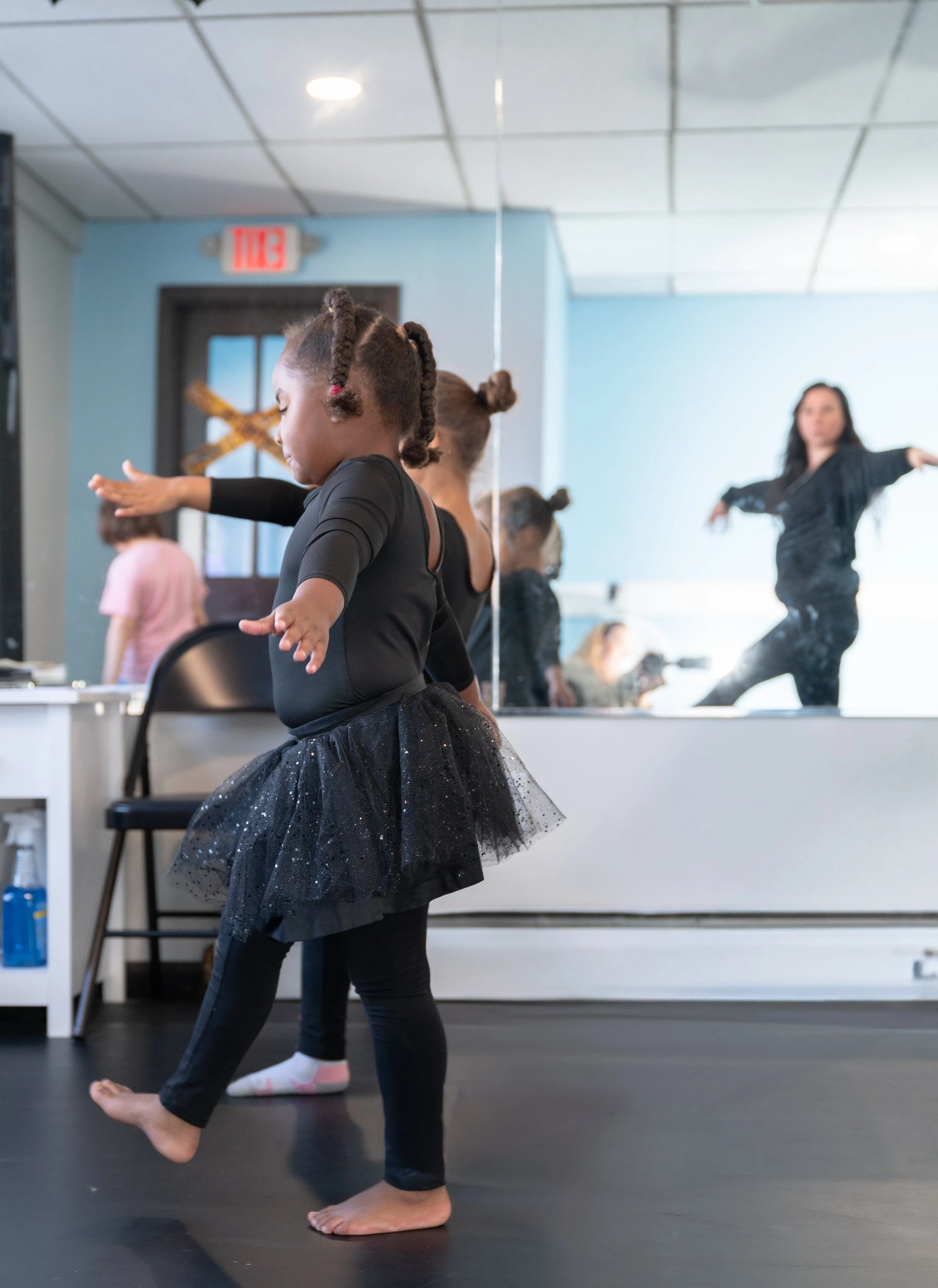 Young girl practicing ballet in dance studio, wearing black tutu and leggings, near mirror reflecting other dancers and instructor.